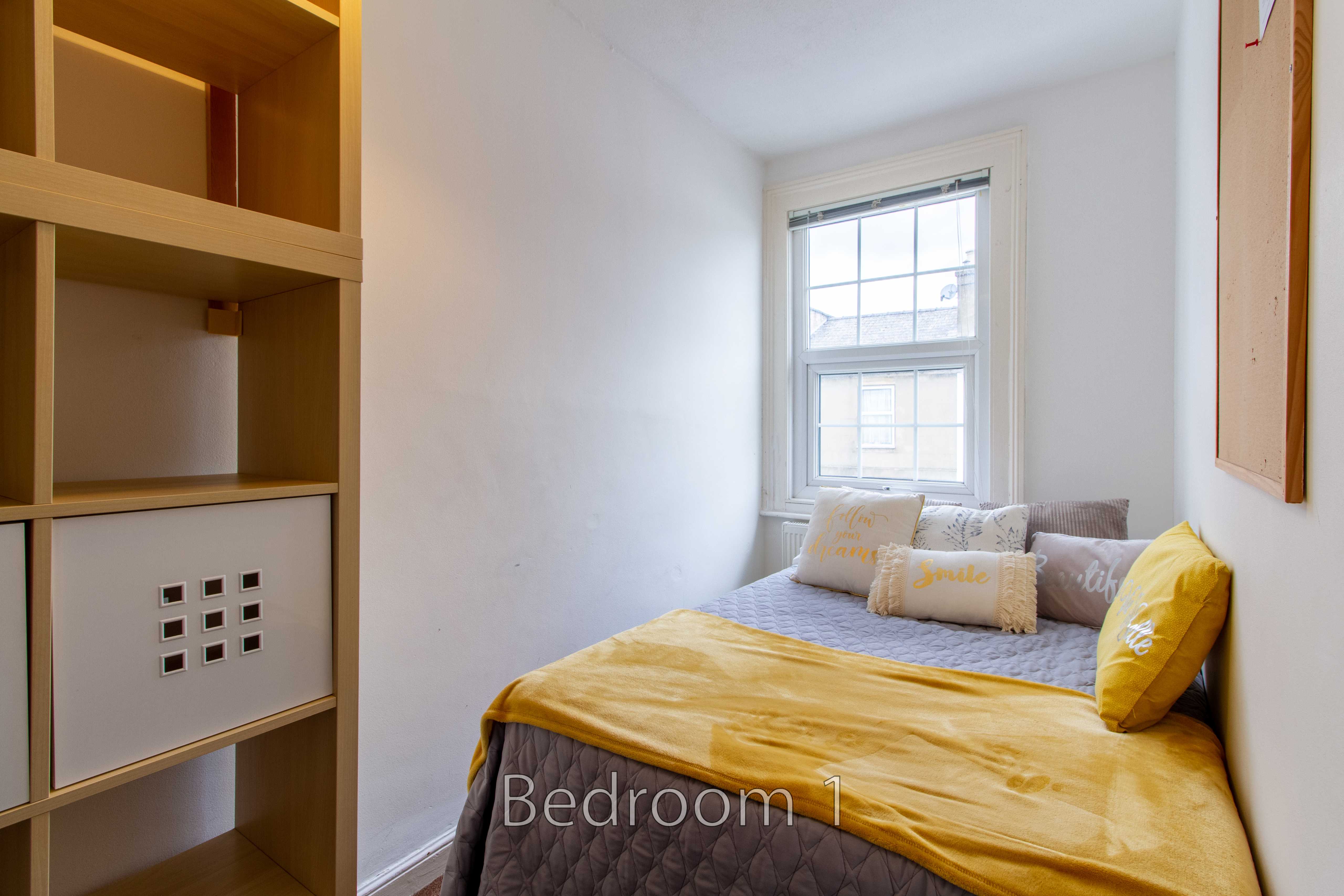 A small bedroom with a double bed, yellow bedding, a window, and built-in shelving.