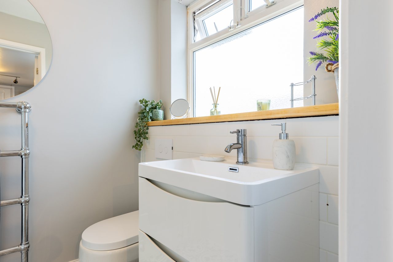 A bright bathroom with a white sink vanity, toilet, towel radiator, wall mirror, tiled backsplash and a window.