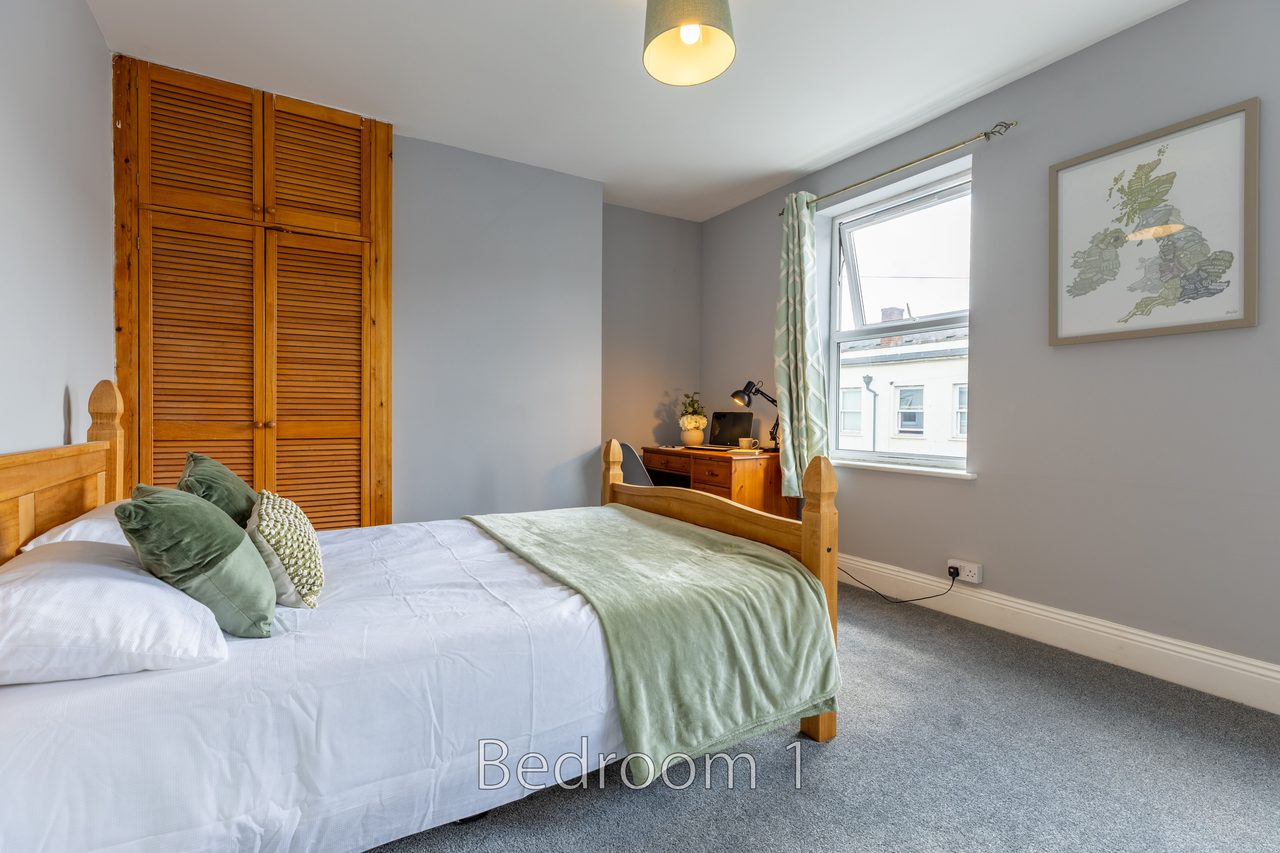 A furnished bedroom with a double bed, wooden wardrobe, window, bedside furniture and framed wall art.