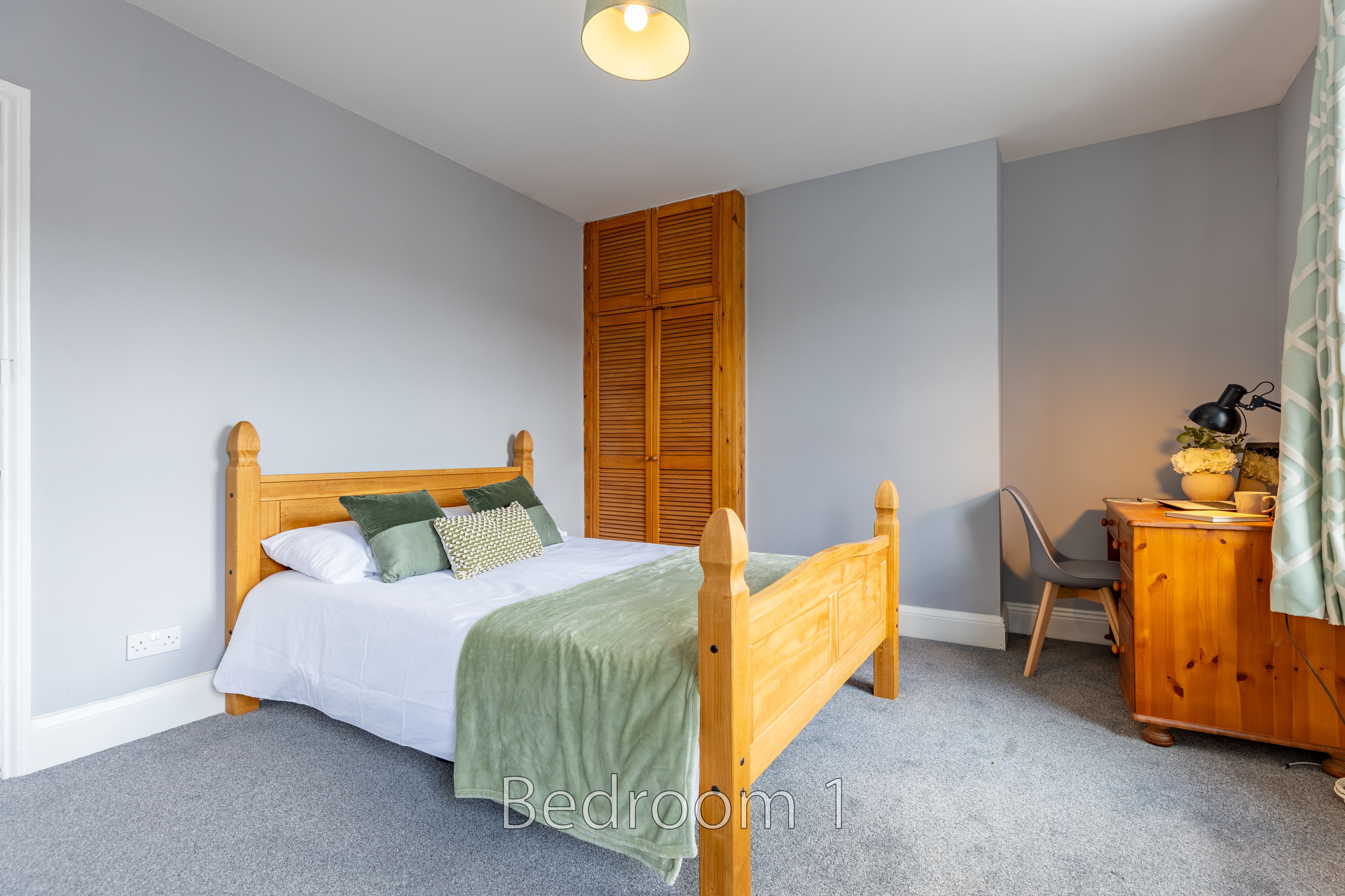 A furnished bedroom with a wooden bed, green and white bedding, grey walls, carpet, a wooden cabinet and a desk.
