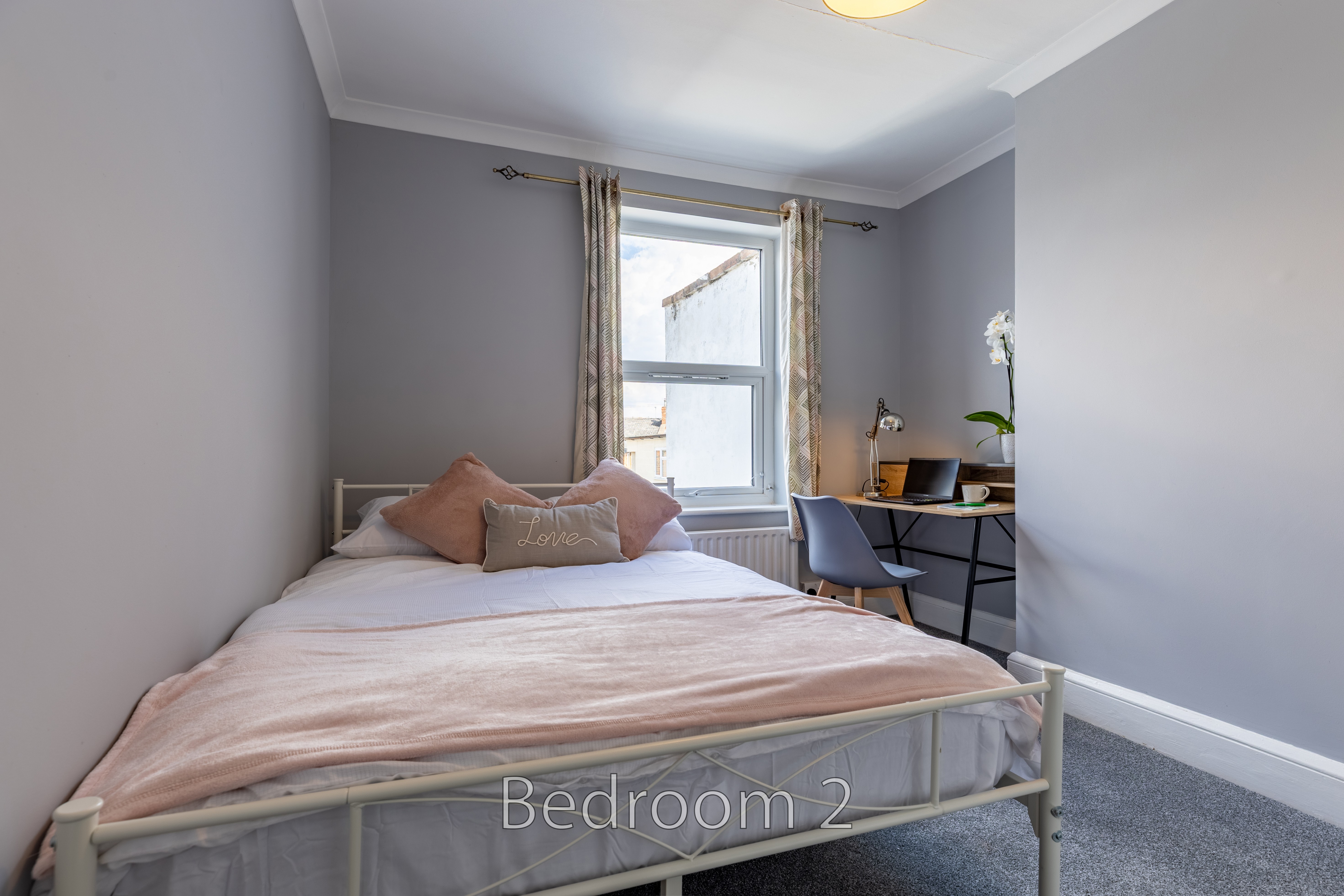 A furnished bedroom with a double bed, desk, chair, window and grey carpet.