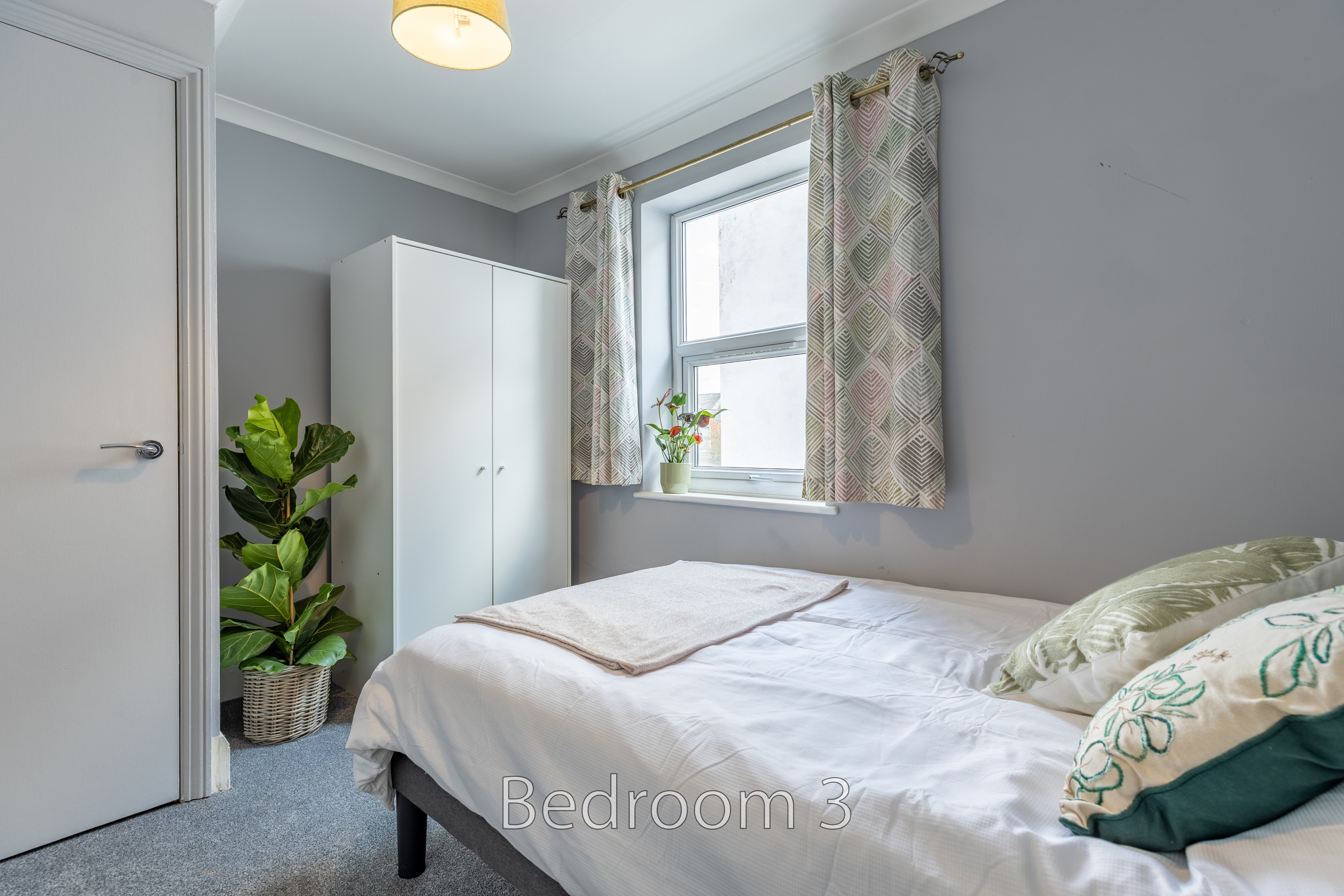 A furnished bedroom with a double bed, wardrobe, window with patterned curtains, plant and carpeted floor.