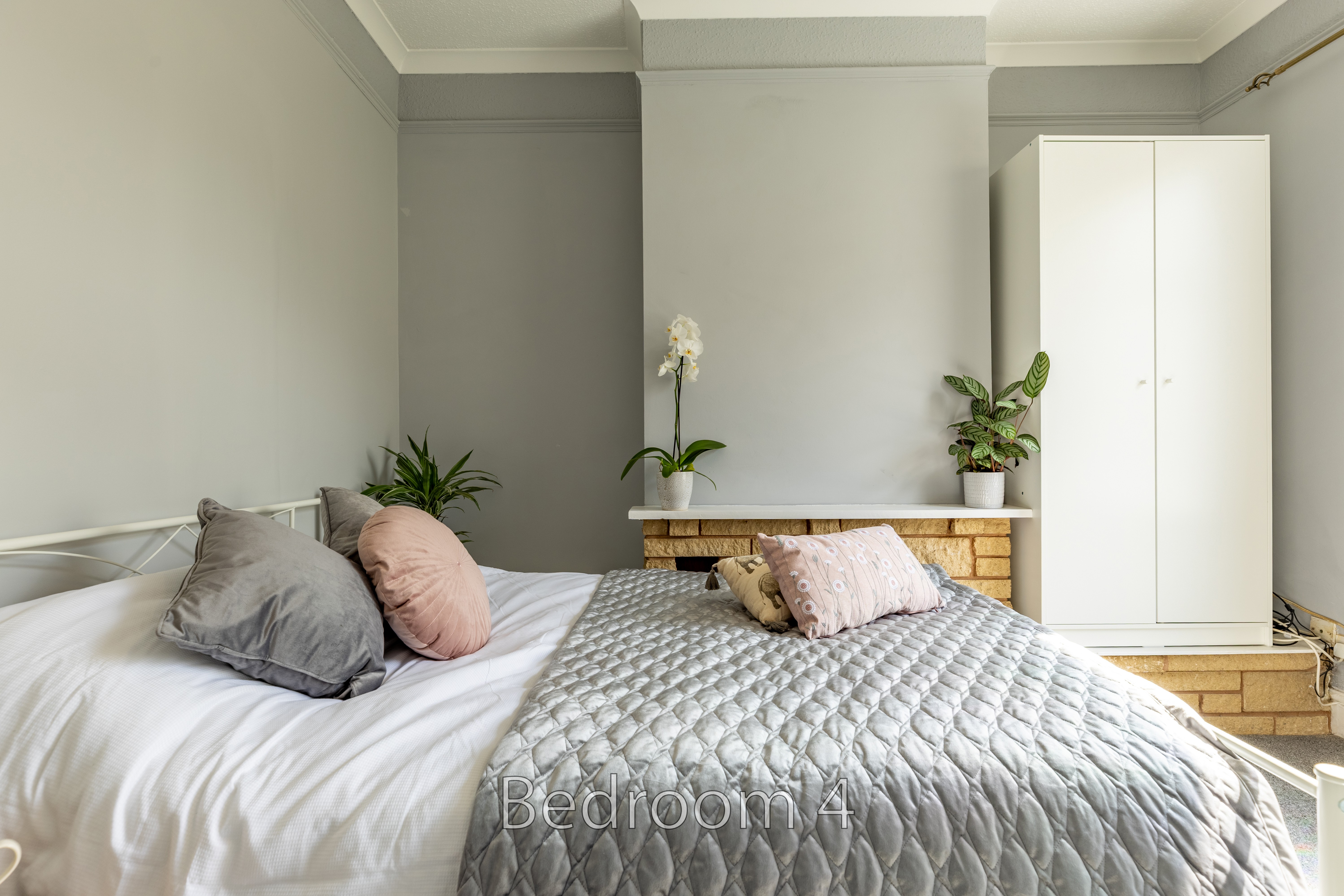 A bedroom with two beds, pillows, a quilted bedspread, plants, and a white wardrobe.
