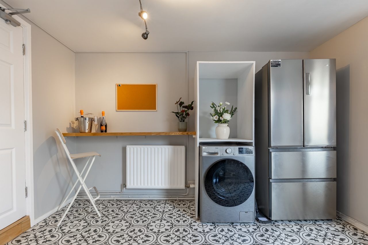 A compact utility area with a washing machine, tall refrigerator, countertop, radiator, folding chair and patterned floor tiles.