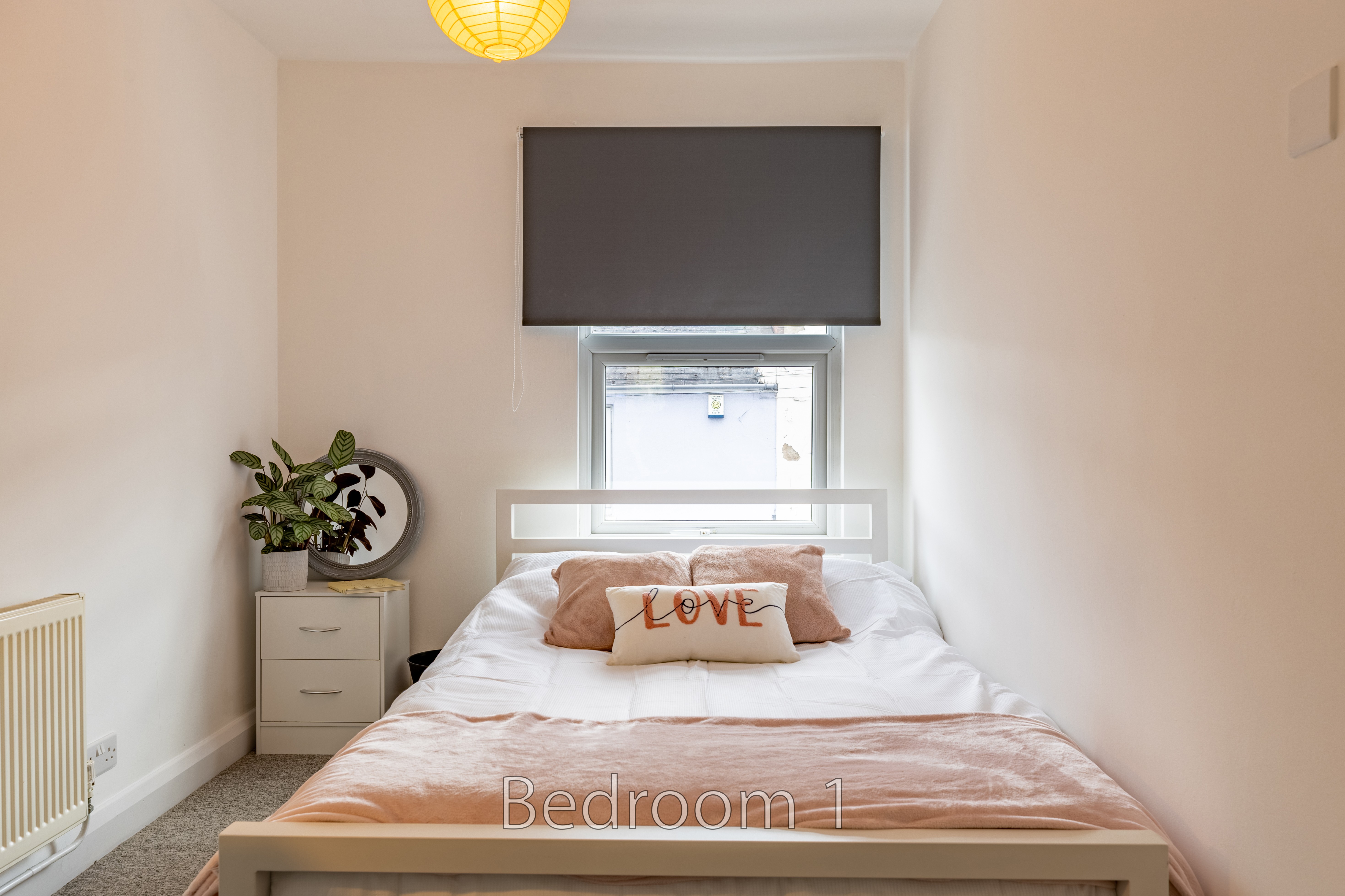 A furnished bedroom with a double bed, window, blinds, bedside cabinet and radiator.