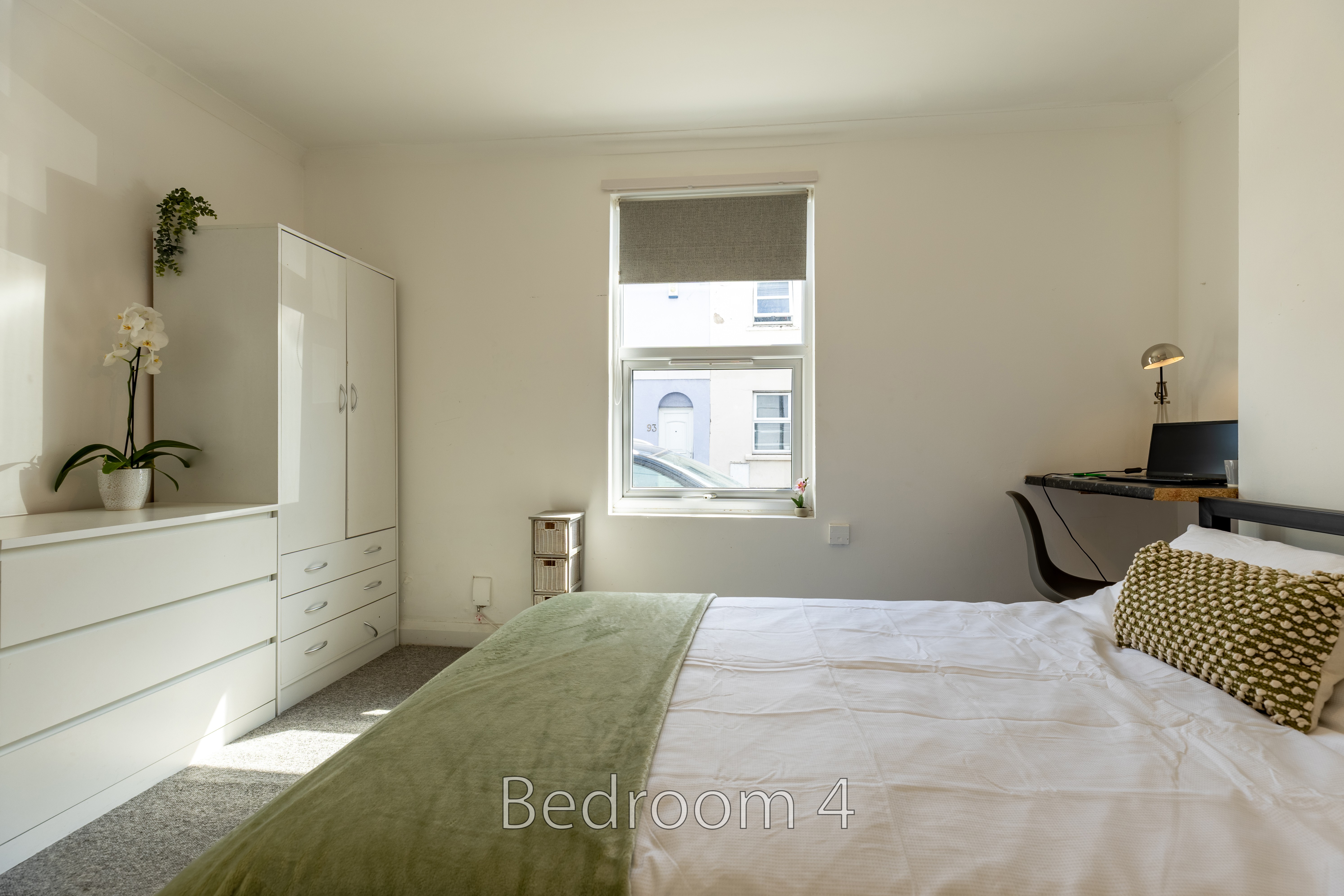 A bright bedroom with a bed, white wardrobes, a desk, a window and potted plants.