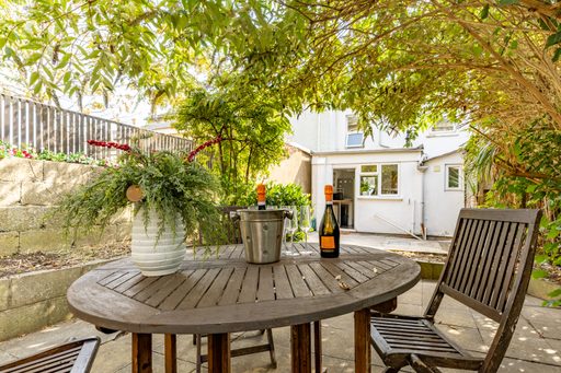 Garden patio with a round wooden table, chairs, plants and a white house in the background.