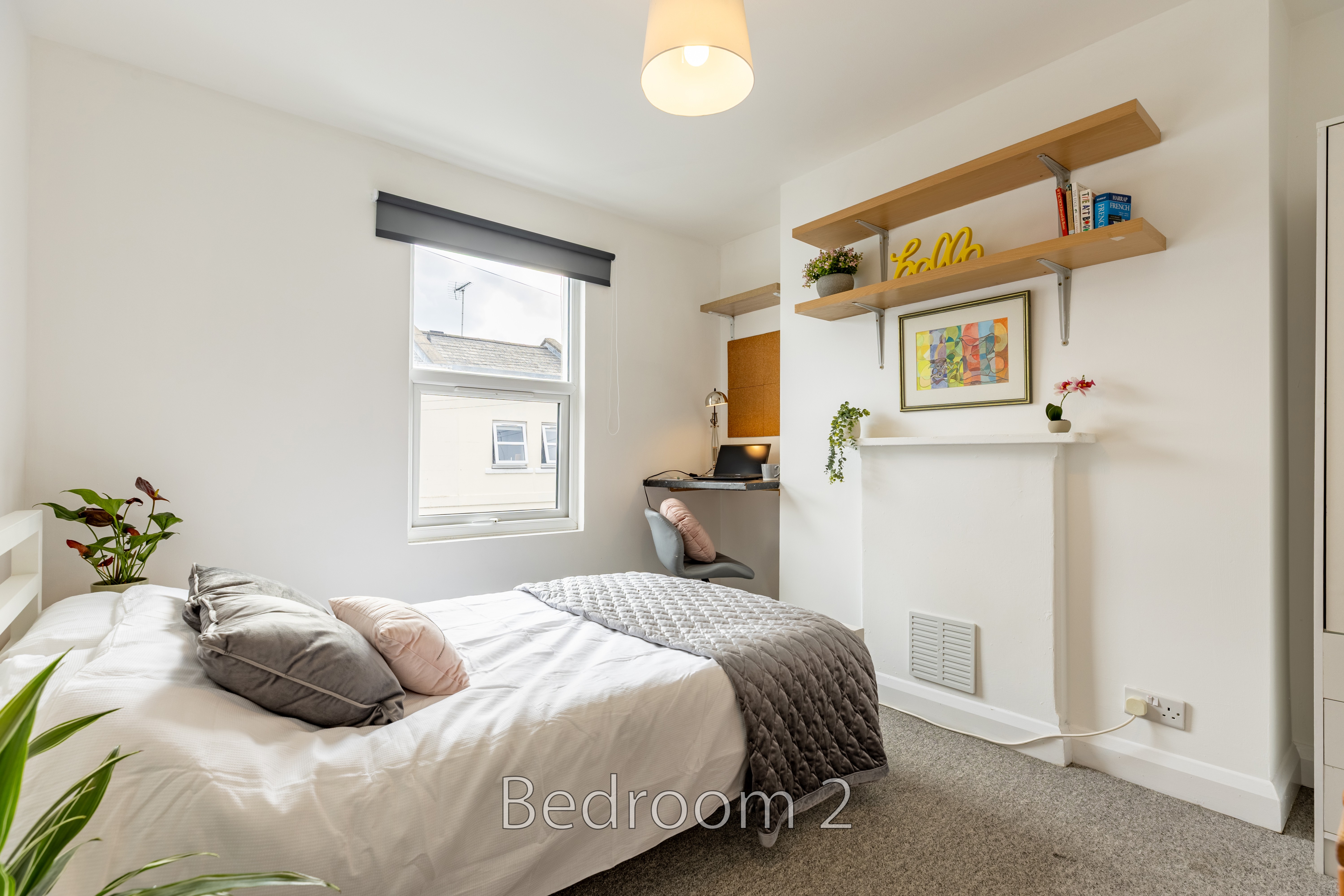 A bright bedroom with a double bed, window, wall shelves, plants and framed artwork.