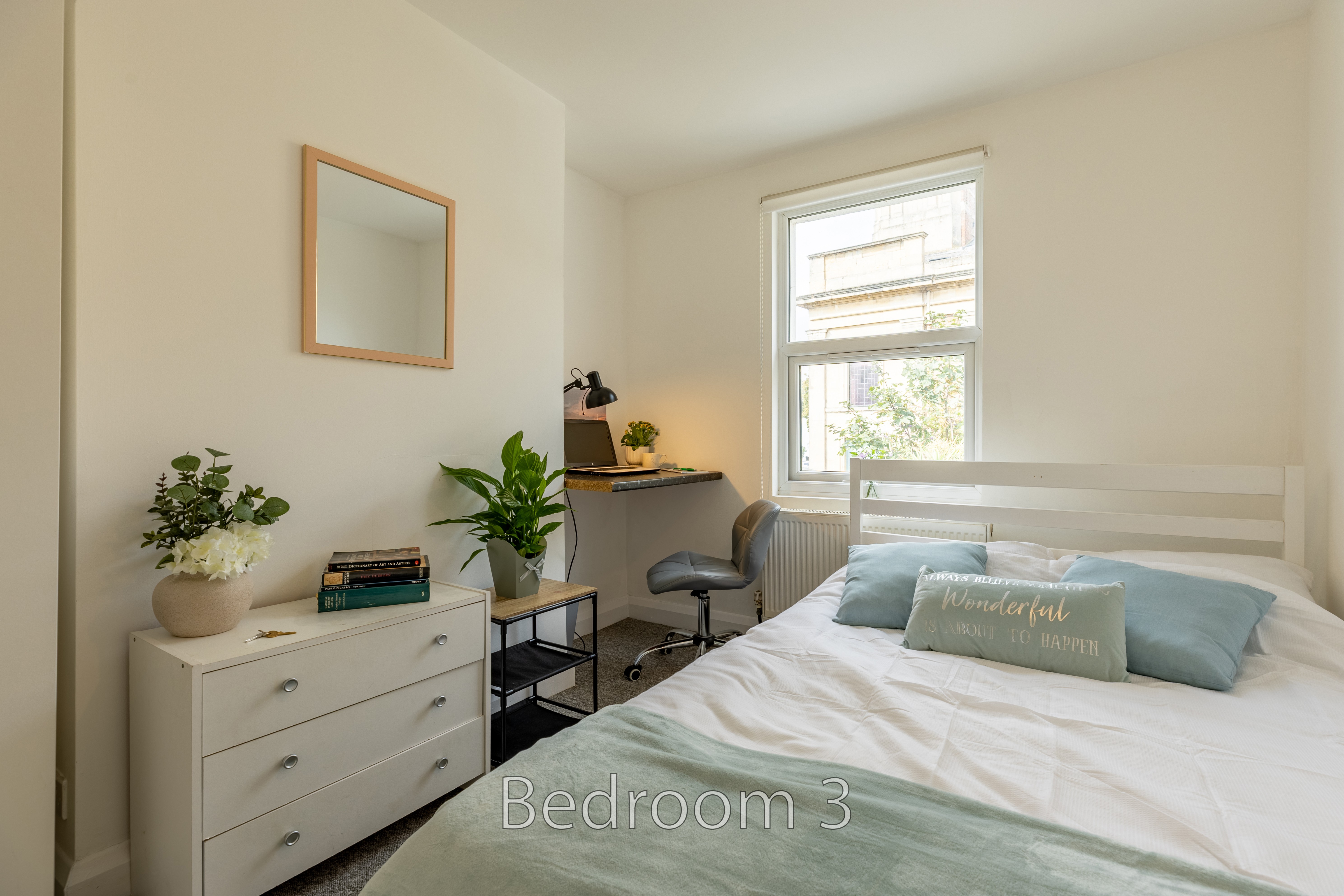 A furnished bedroom with a double bed, dresser, mirror, plants, desk and window.