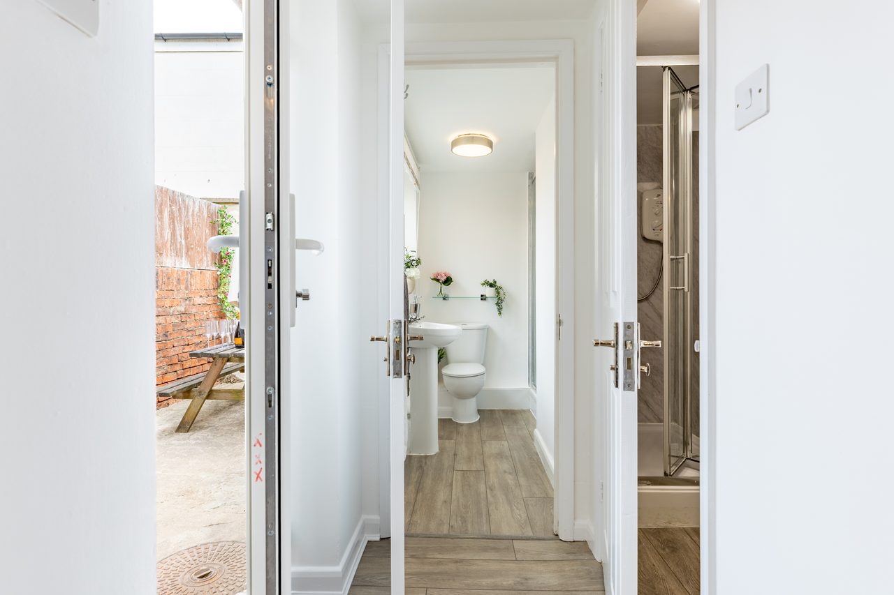 Bright hallway with several open doors, showing a toilet and a shower room, with an exterior doorway on the left.