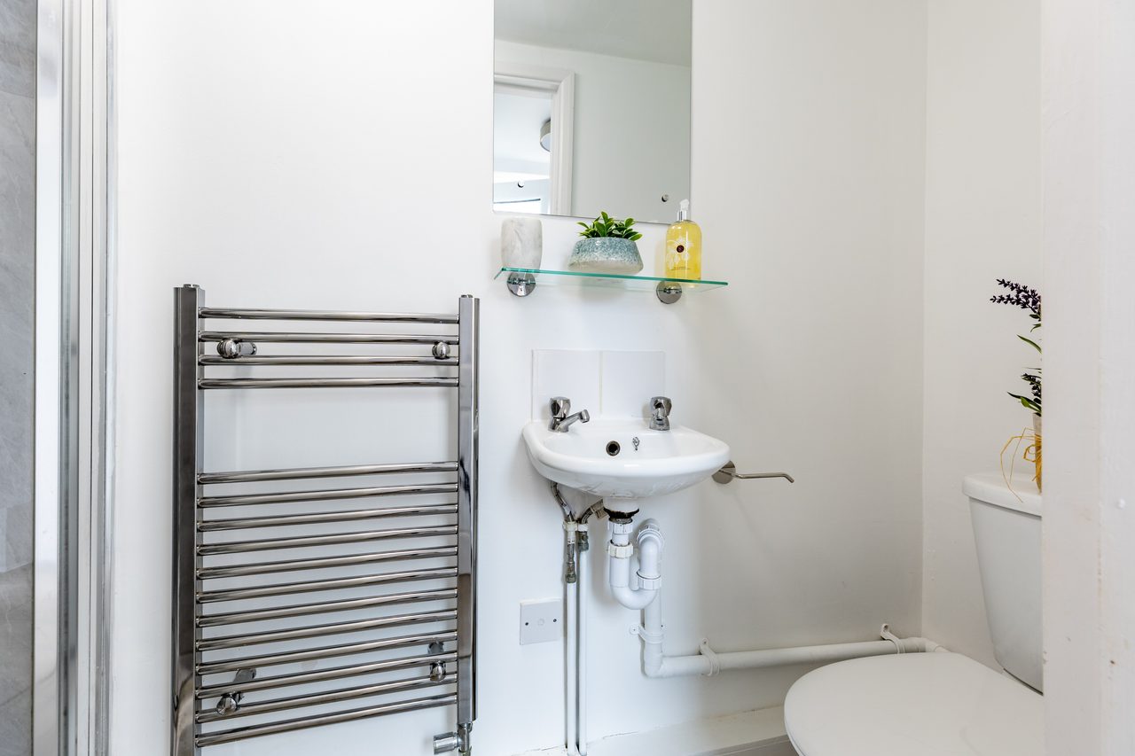 Small bathroom with a wall-mounted sink, chrome heated towel rail, toilet and shelf with toiletries.