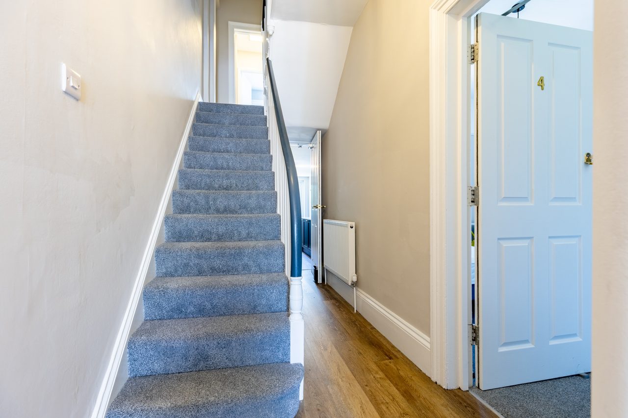 A shared hallway with a carpeted staircase, wooden flooring, a radiator, and an open numbered door.