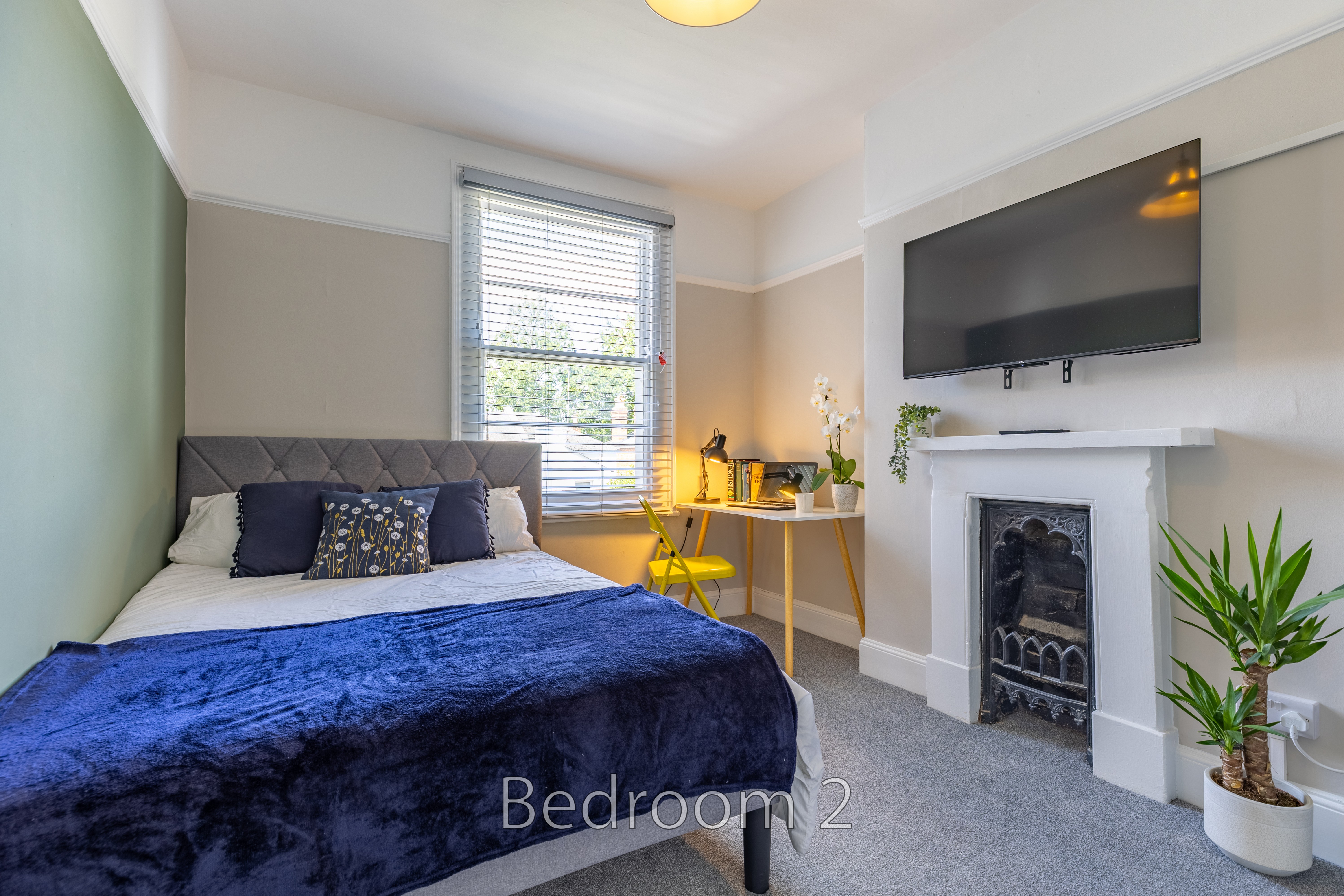 A furnished bedroom with a double bed, television above a fireplace, window, desk and potted plant.