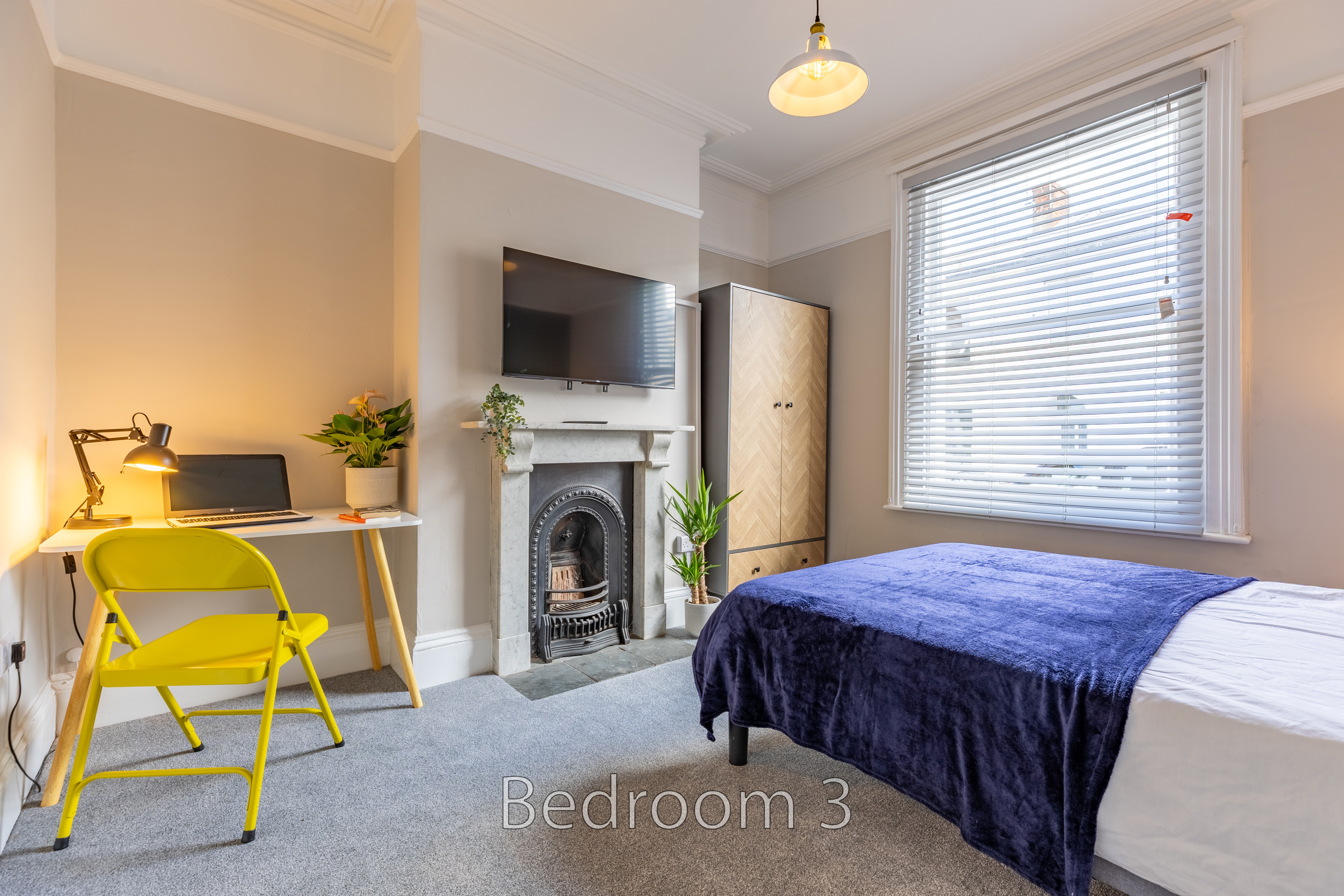 A bedroom with a bed, fireplace, desk, chair, wardrobe and window with blinds.