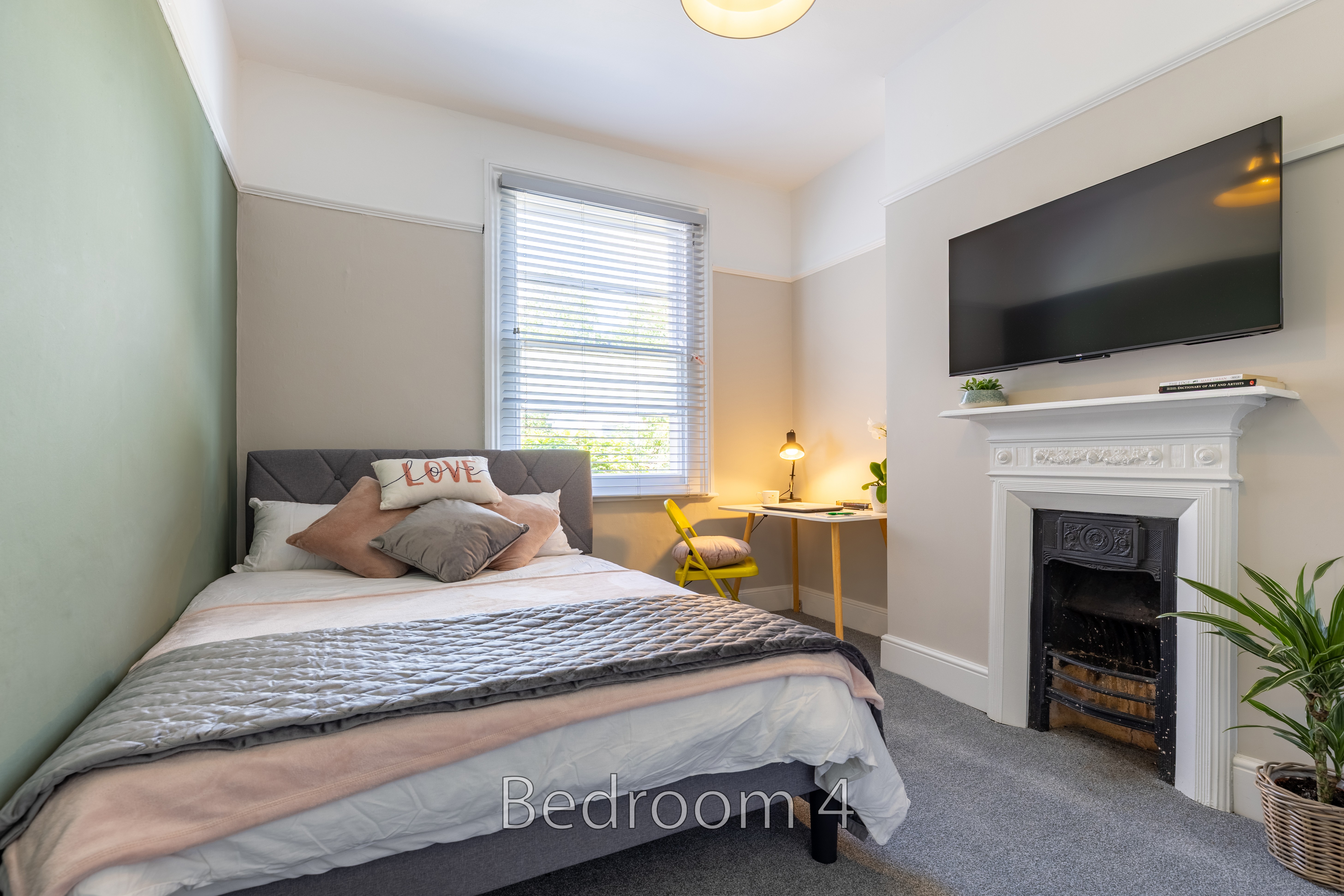 A furnished bedroom with a double bed, wall-mounted television, fireplace, window and armchair.
