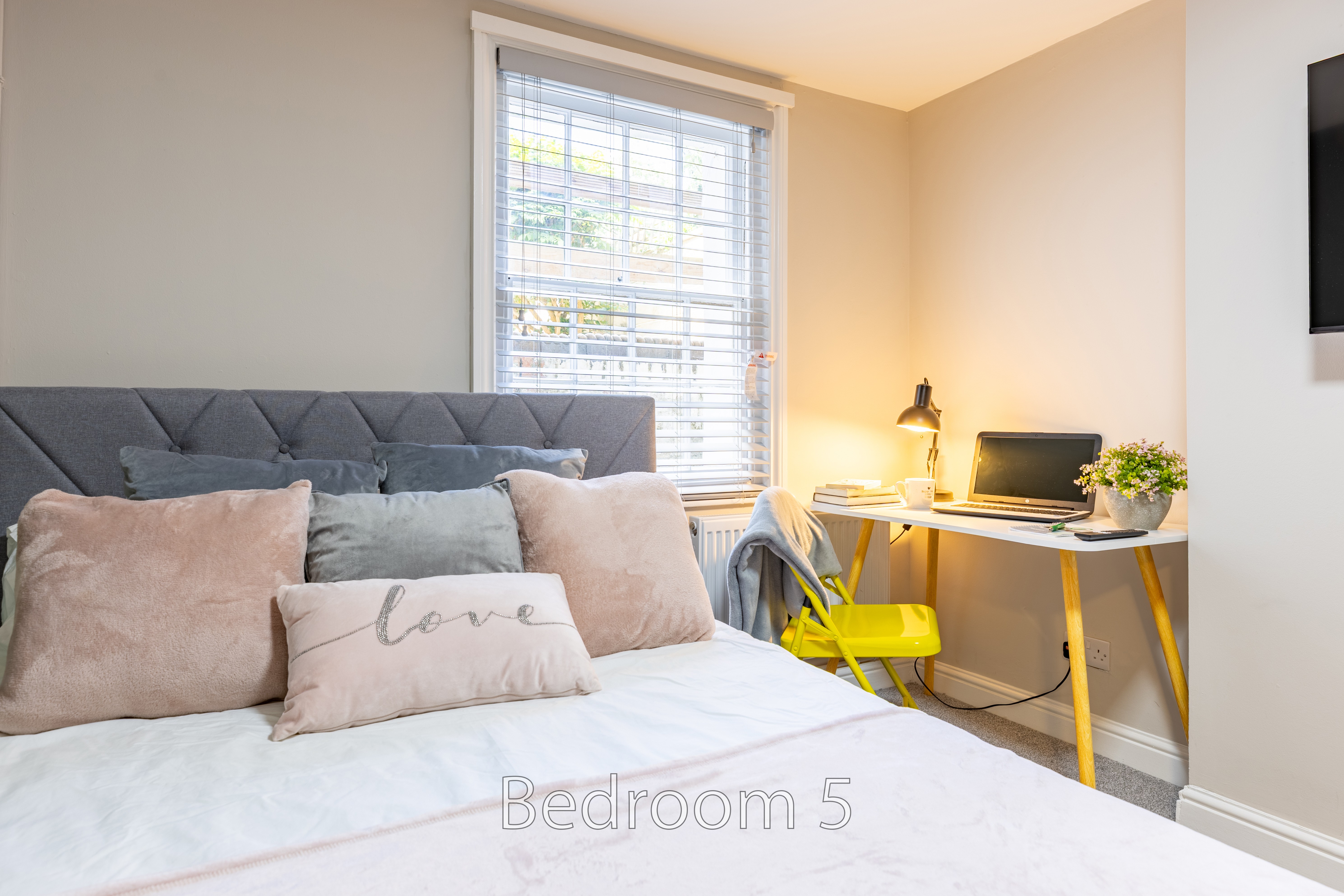 A furnished bedroom with a bed, desk, chair, window and wall-mounted television.