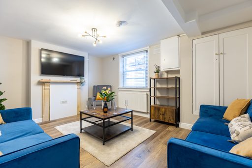 Bright furnished living room with blue sofas, a television, coffee table, shelving, and built-in cupboards.