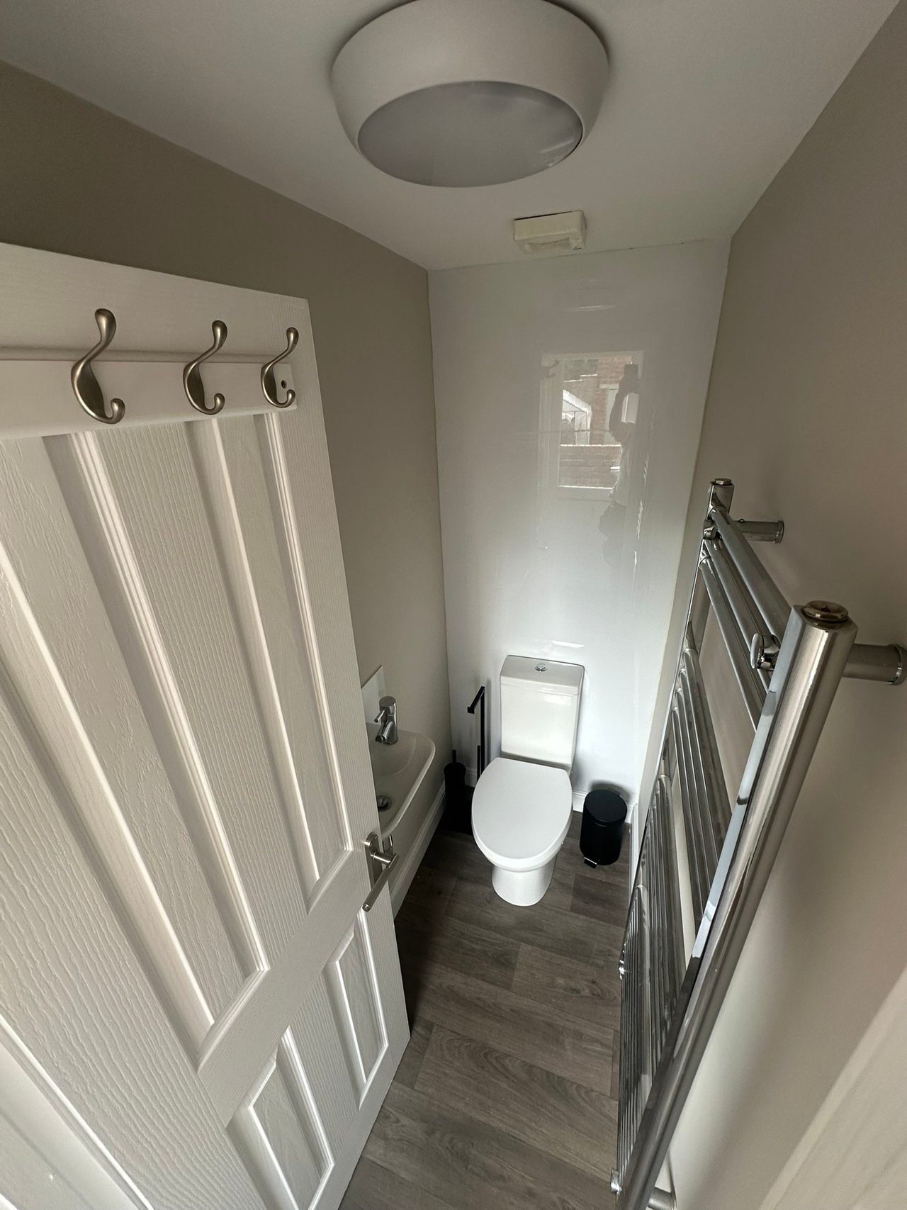 A narrow toilet room with a white toilet, dark wood-effect flooring, a radiator, wall hooks and a metal stair railing.