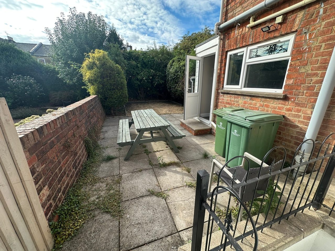 A paved backyard patio with a picnic table, brick walls, garden plants, bins and a house entrance.