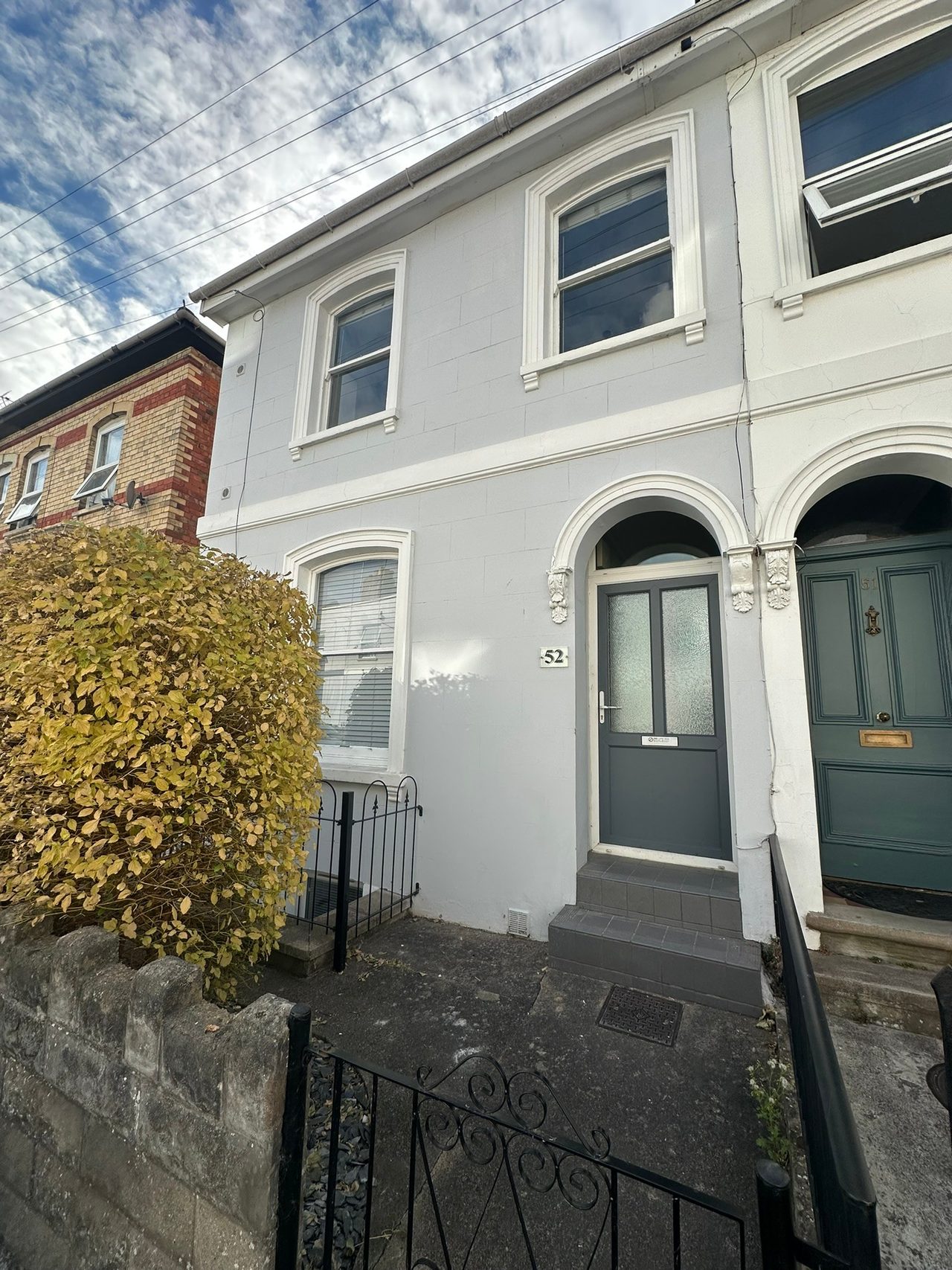 Front exterior of a grey terraced house with a dark entrance door, upper windows, railings and a hedge.