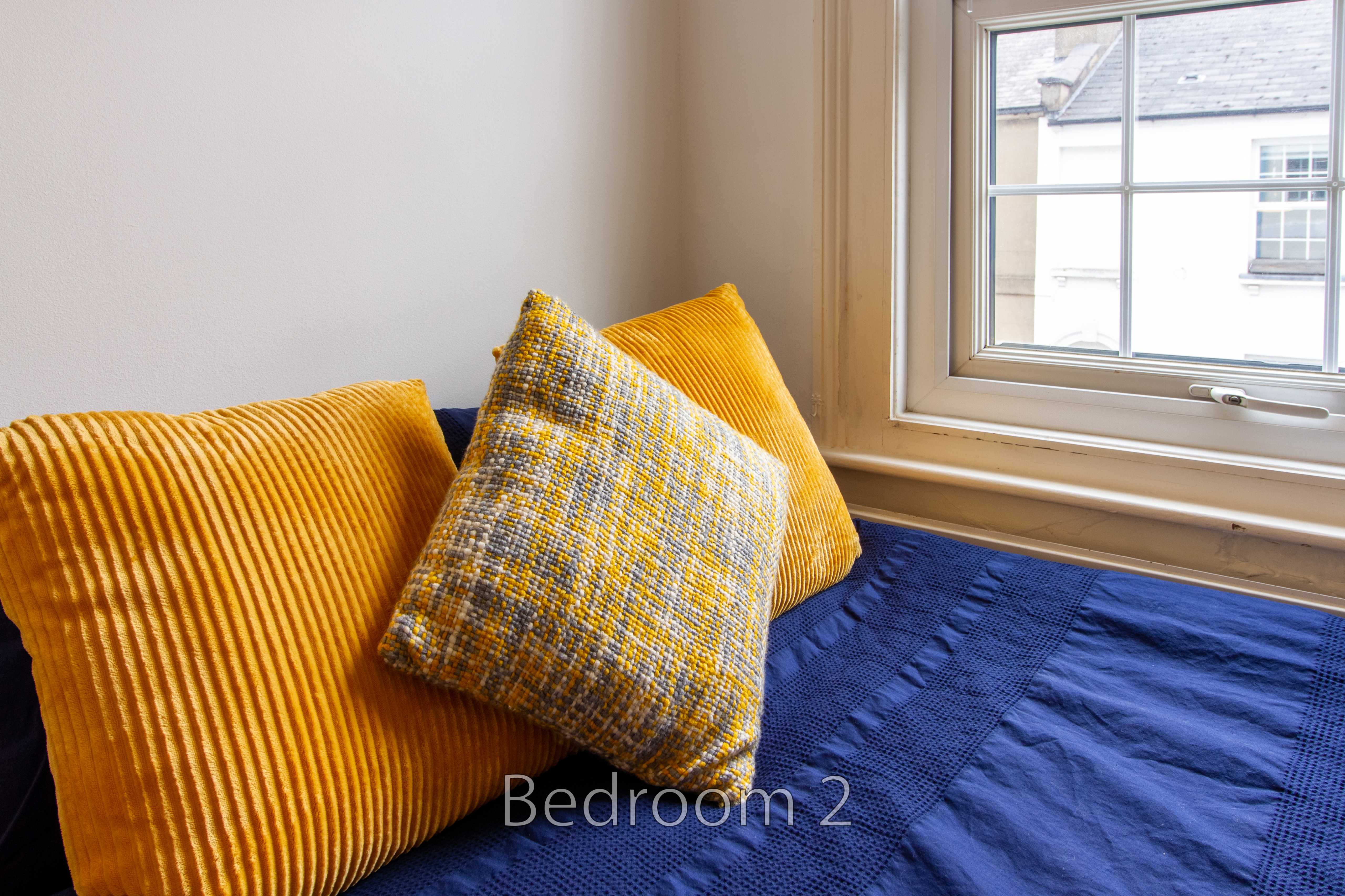 A bed with blue bedding and mustard and patterned pillows beside a window.
