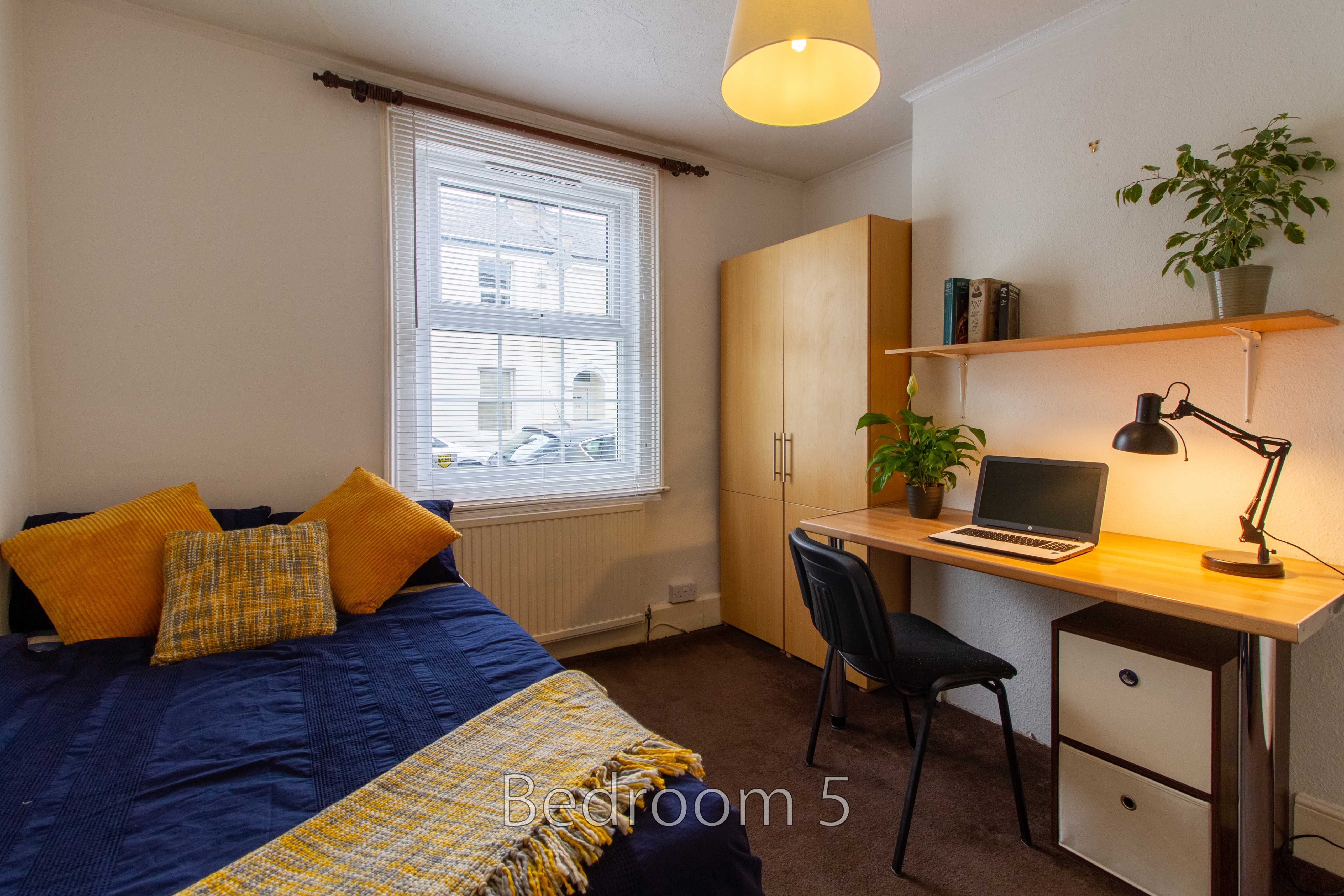 A furnished bedroom with a single bed, wardrobe, desk, chair, window and plants.
