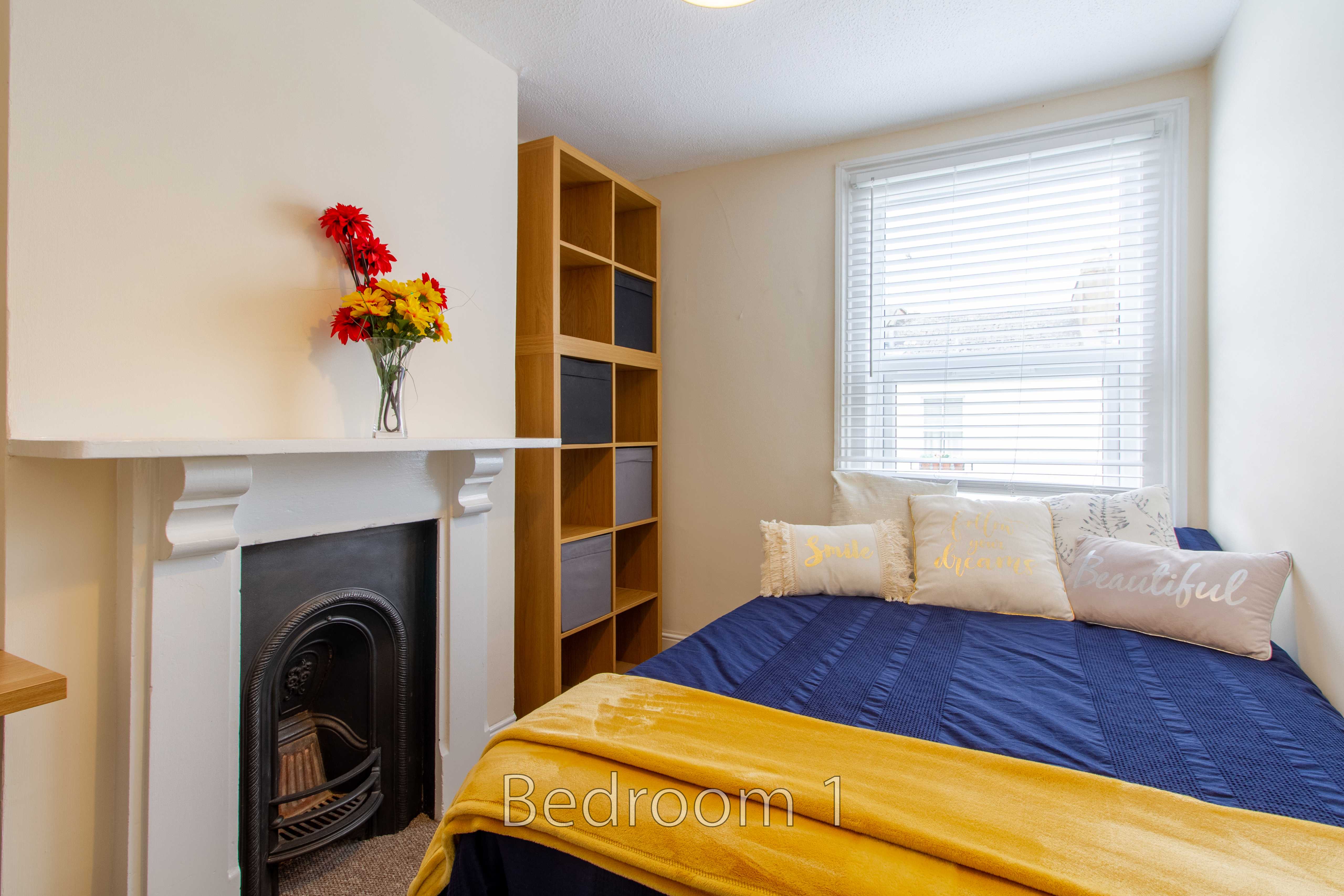 A bedroom with a bed, blue and mustard bedding, a fireplace, shelving, and a window with blinds.