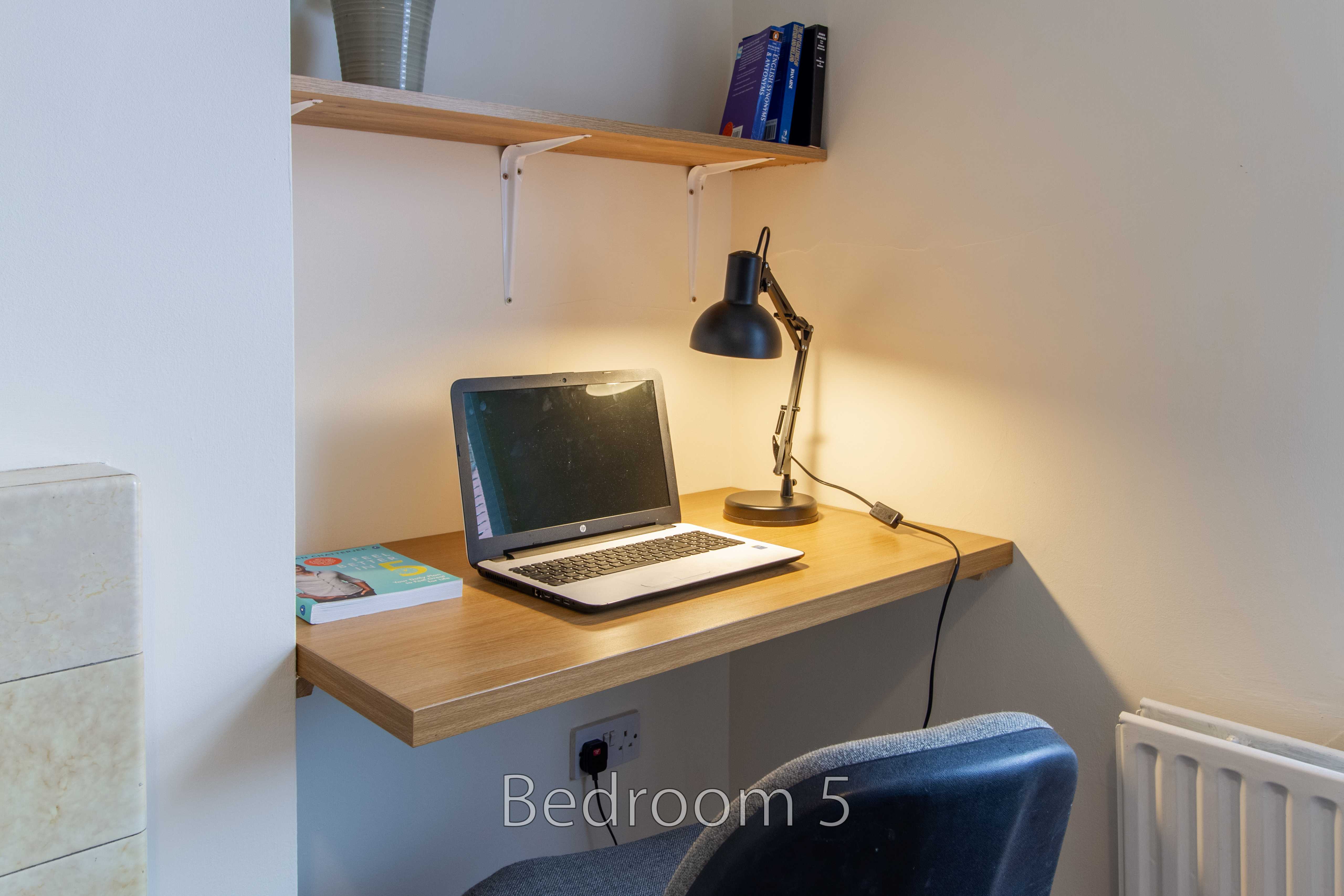 A compact bedroom workspace with a desk, laptop, lamp, chair, shelves and radiator.