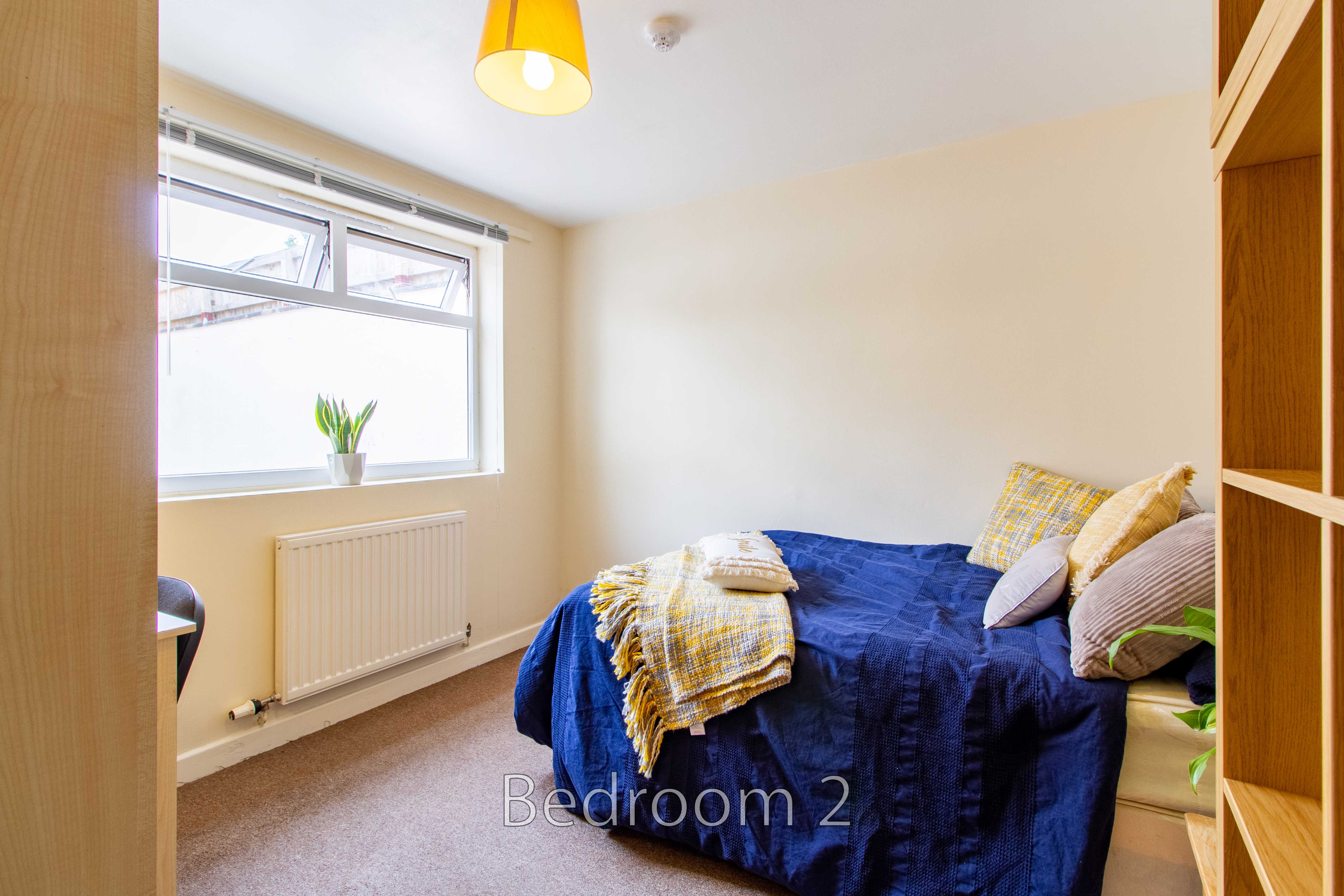 A furnished bedroom with a double bed, patterned cushions, a window, radiator and open wooden shelving.