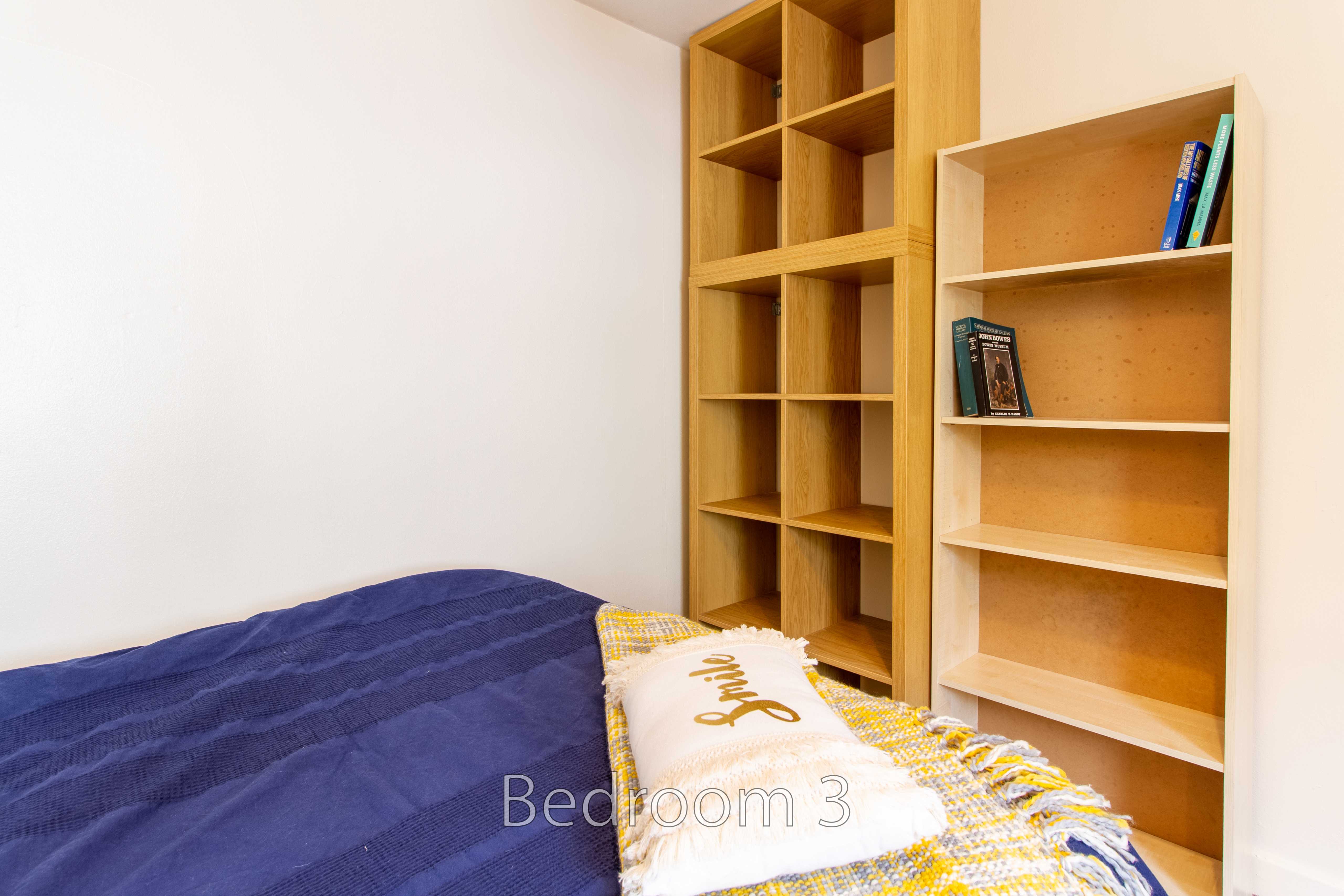 A bedroom with a bed covered by blue and patterned blankets, beside wooden shelving units.