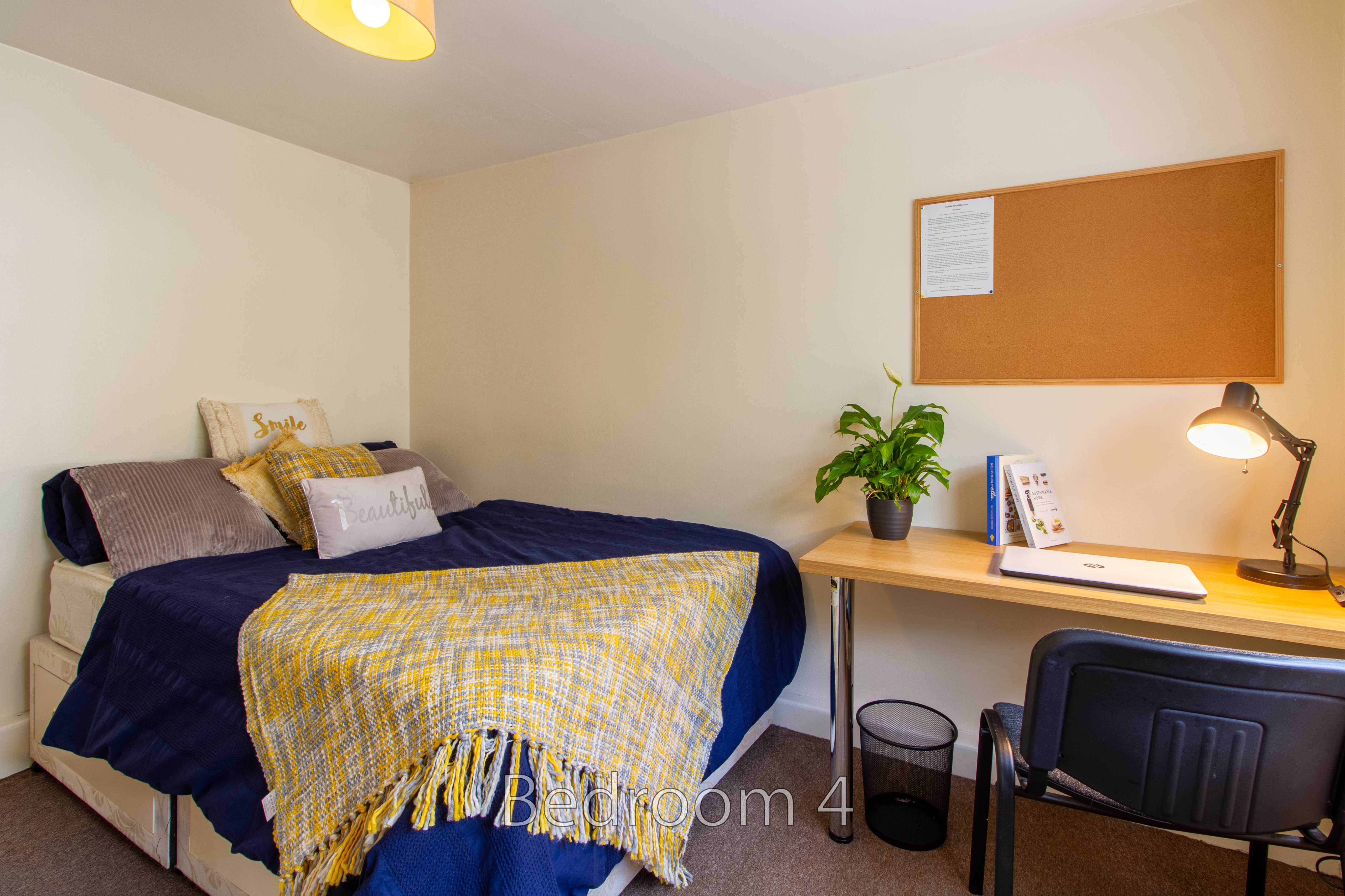 A furnished bedroom with a bed, desk, chair, lamp, plant and wall noticeboard.