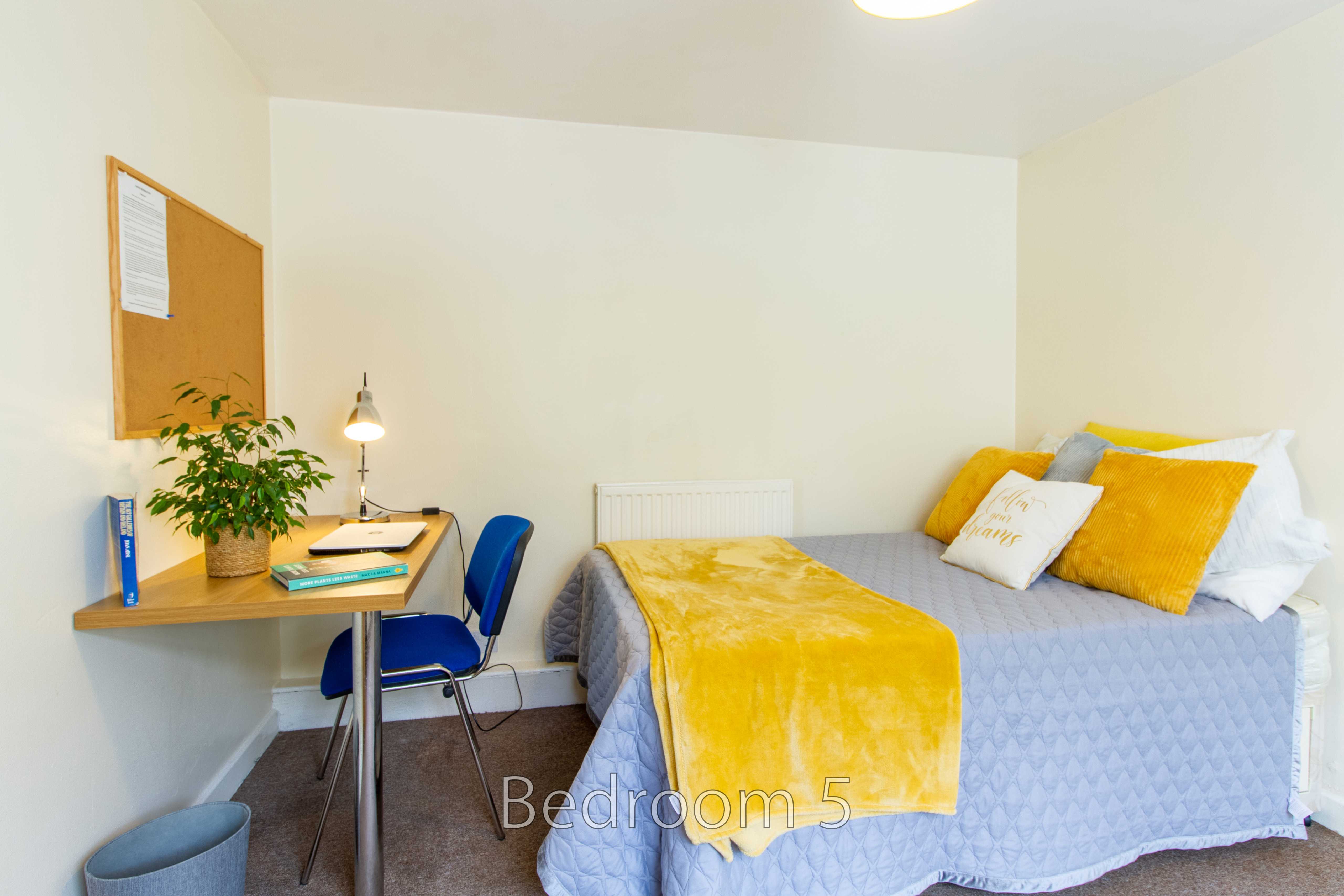 A furnished bedroom with a single bed, yellow and white bedding, a desk, chair, lamp and potted plant.