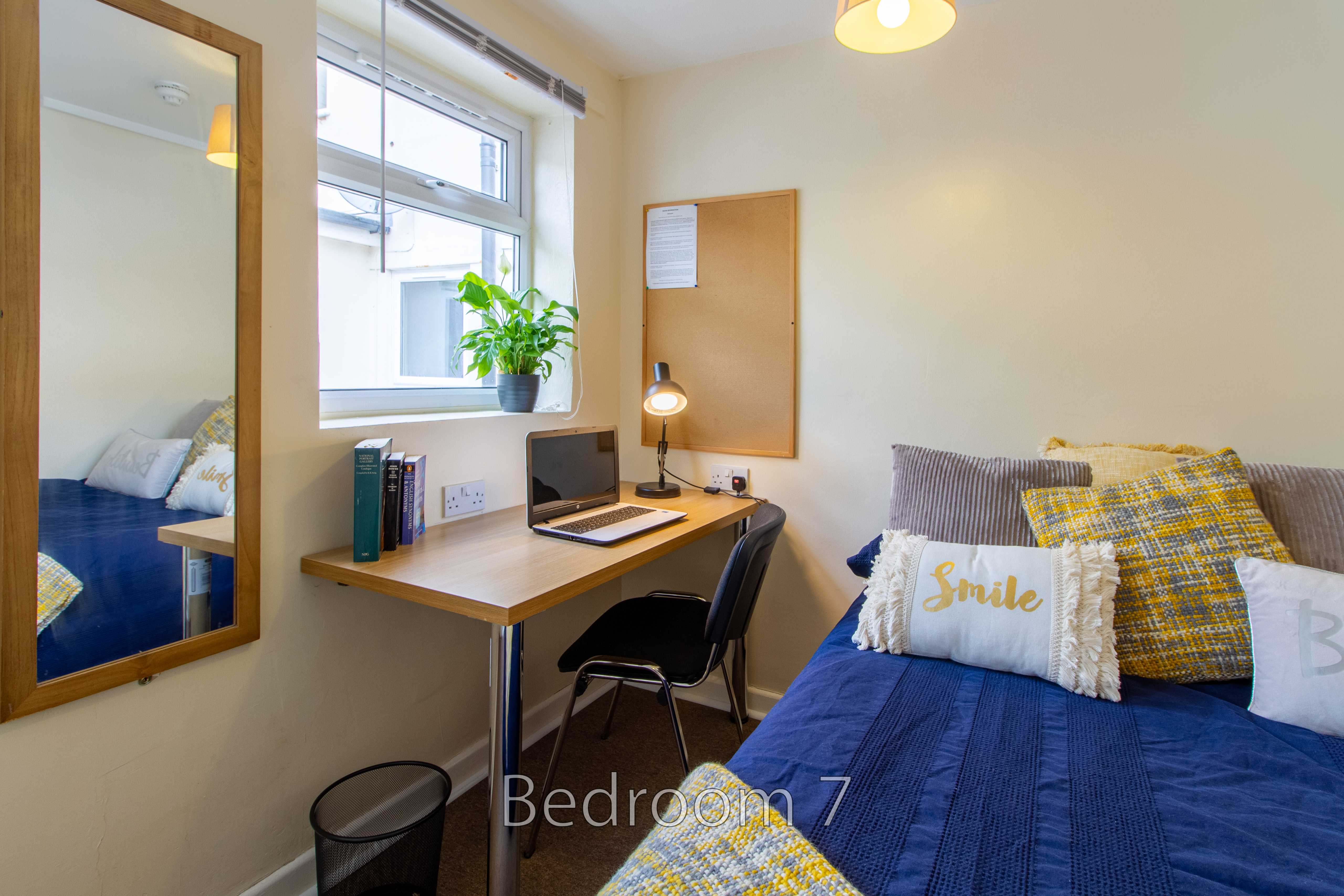 A furnished bedroom with a bed, desk, chair, window, mirror and bedside lamp.