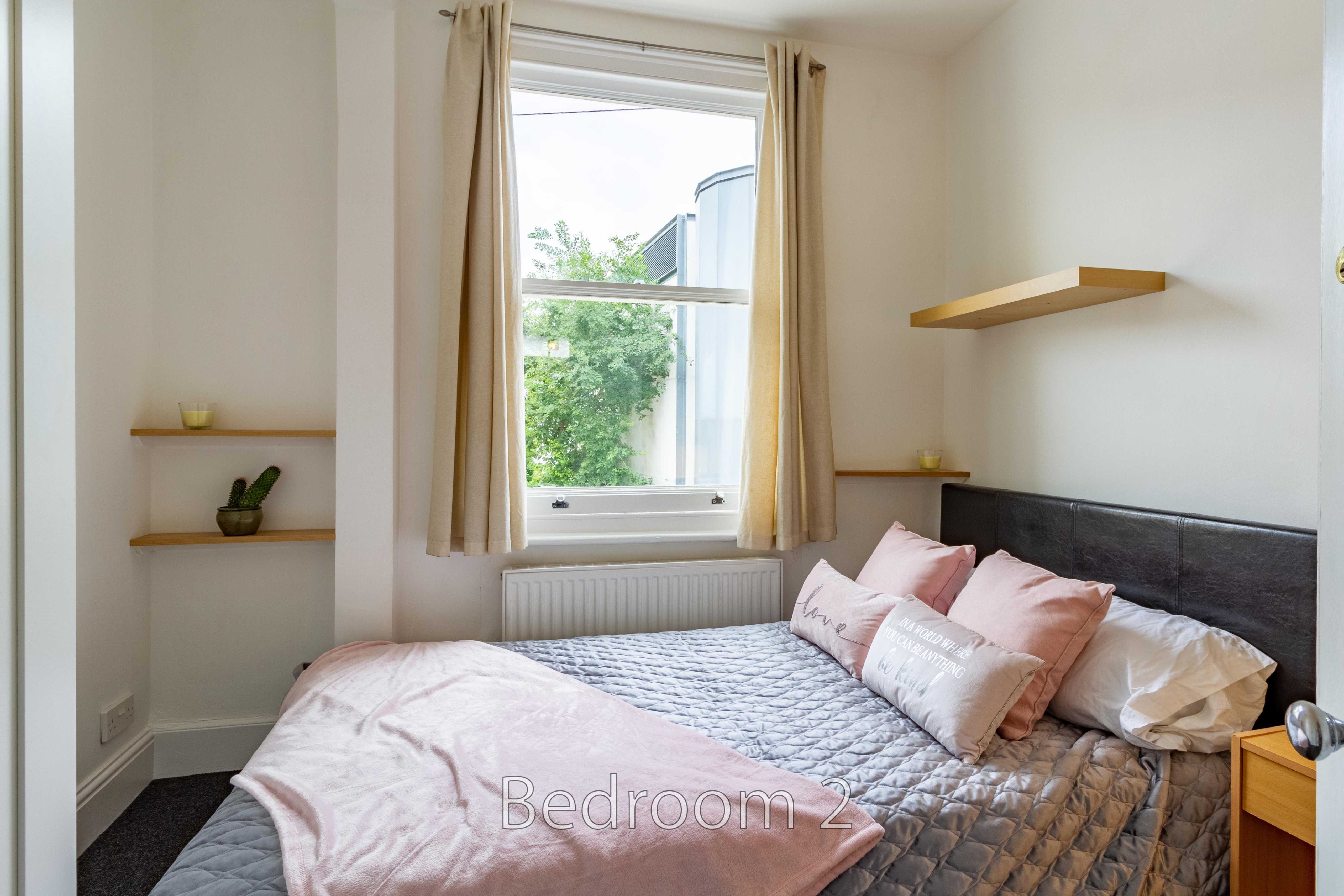 A furnished bedroom with a double bed, window, curtains, shelves and a wall-mounted shelf.
