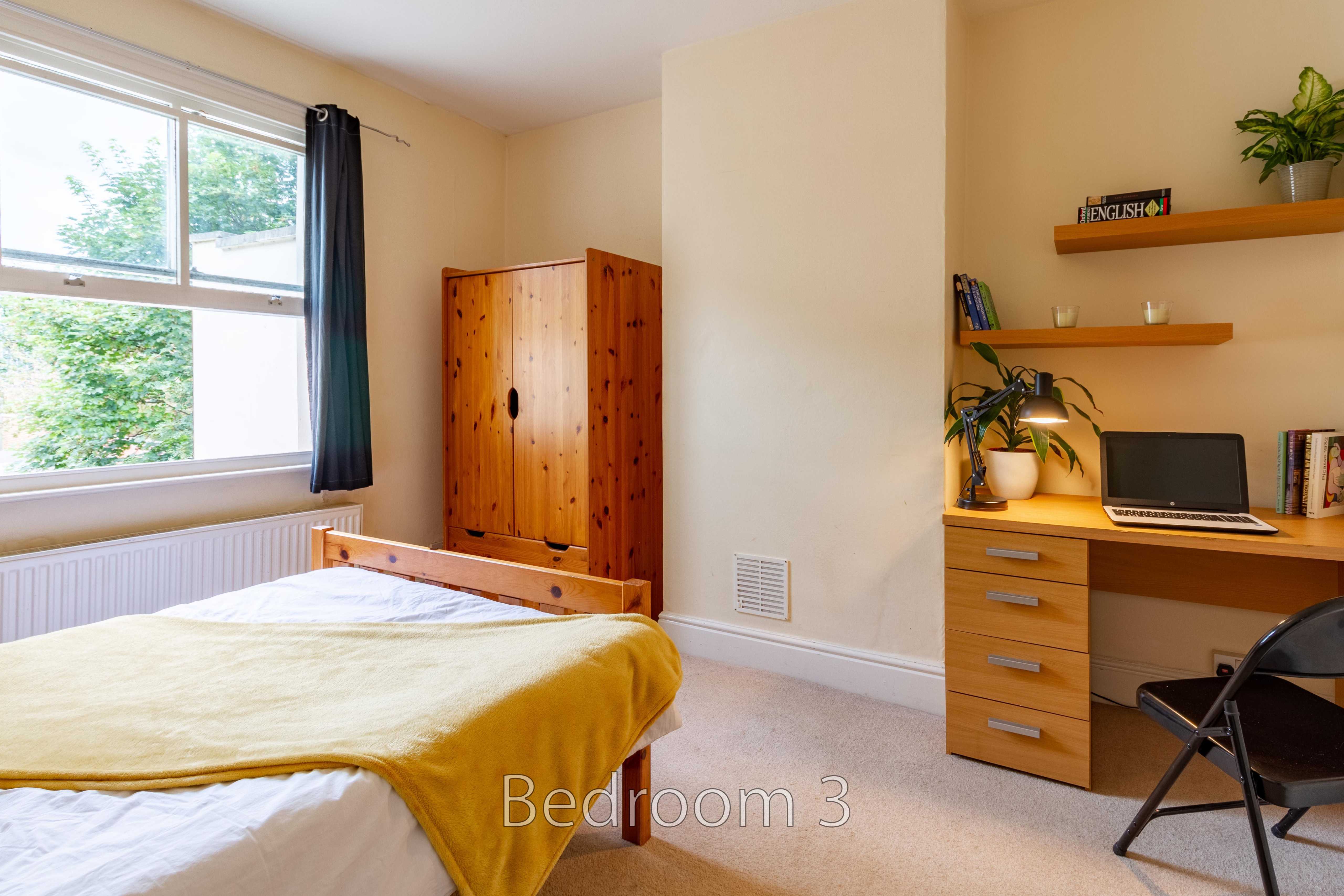A bedroom with a bed, wooden wardrobe, desk, chair, shelves and a large window.