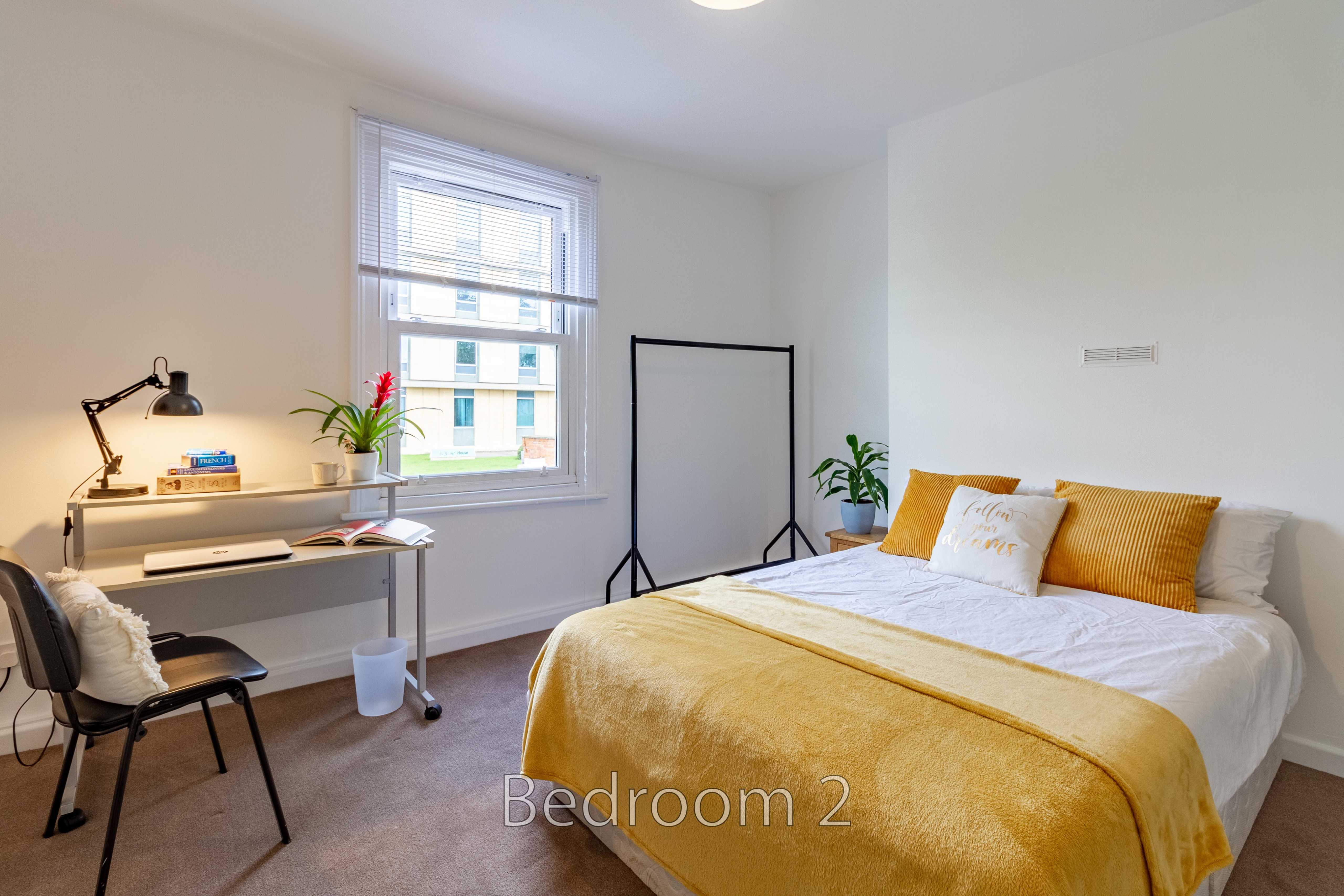 A bright bedroom with a double bed, yellow bedding, desk, chair, window, plants and a clothes rack.