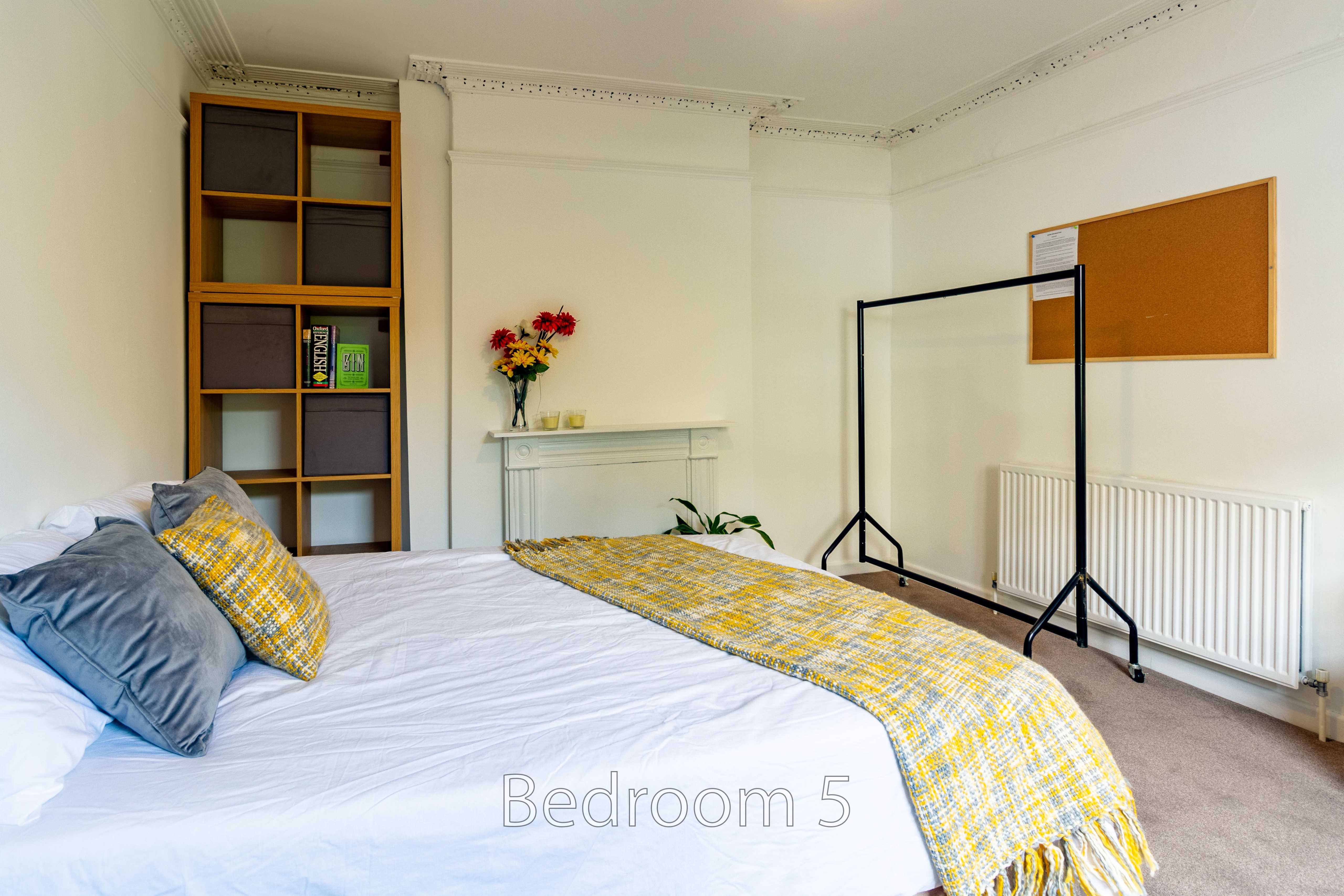 A bedroom with a bed, yellow patterned runner, shelving, fireplace mantel, radiator and window.