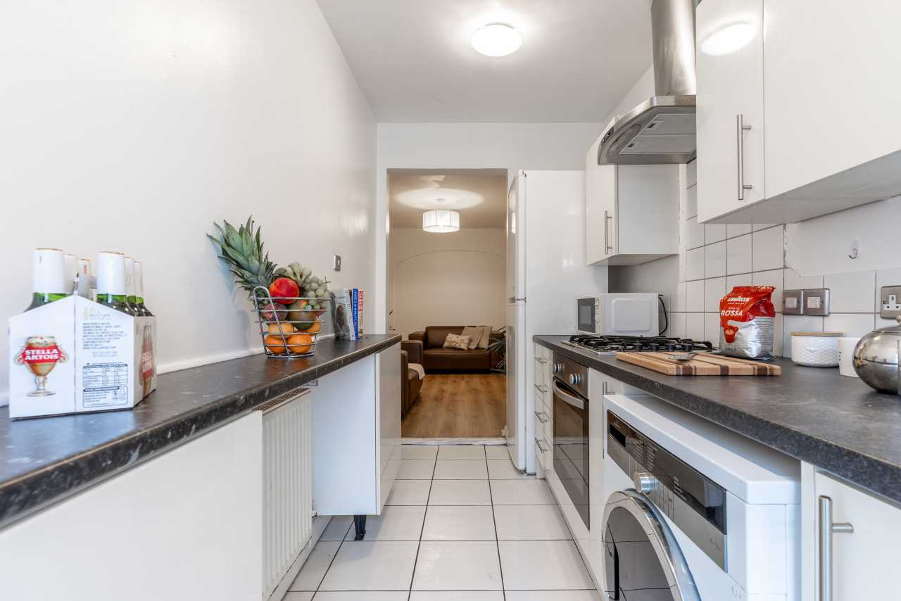 Bright fitted kitchen with white cabinets, dark worktops, tiled floor, cooker, washing machine and a living area visible through the doorway.