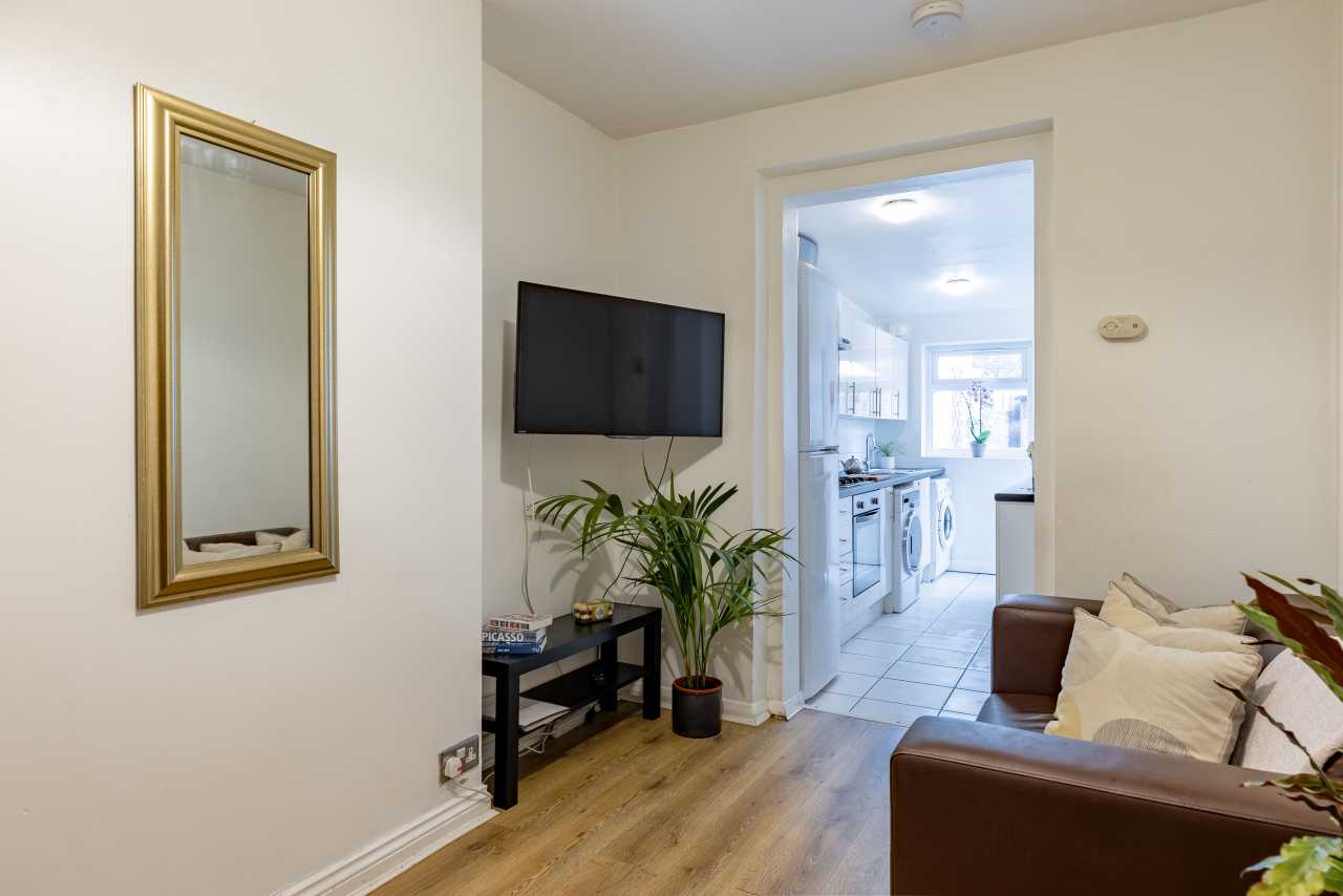 Living room with a wall-mounted television, sofa, mirror and doorway leading to a bright kitchen.