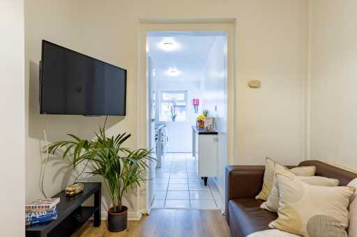 Living room with wall-mounted television, potted plant and sofa, looking through to a bright kitchen.
