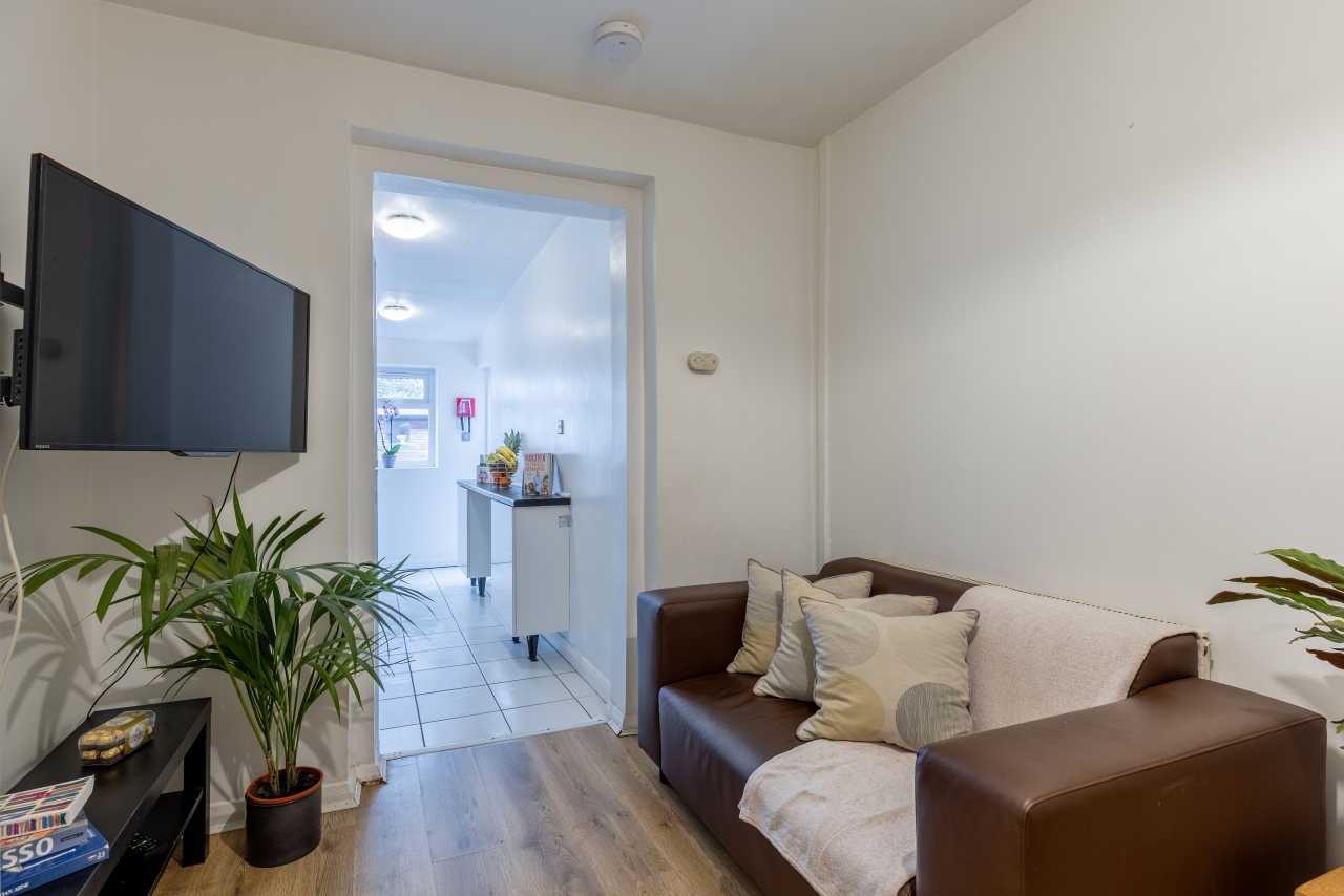 Shared living room with a wall-mounted television, sofa, plants and an open doorway leading to a tiled kitchen area.