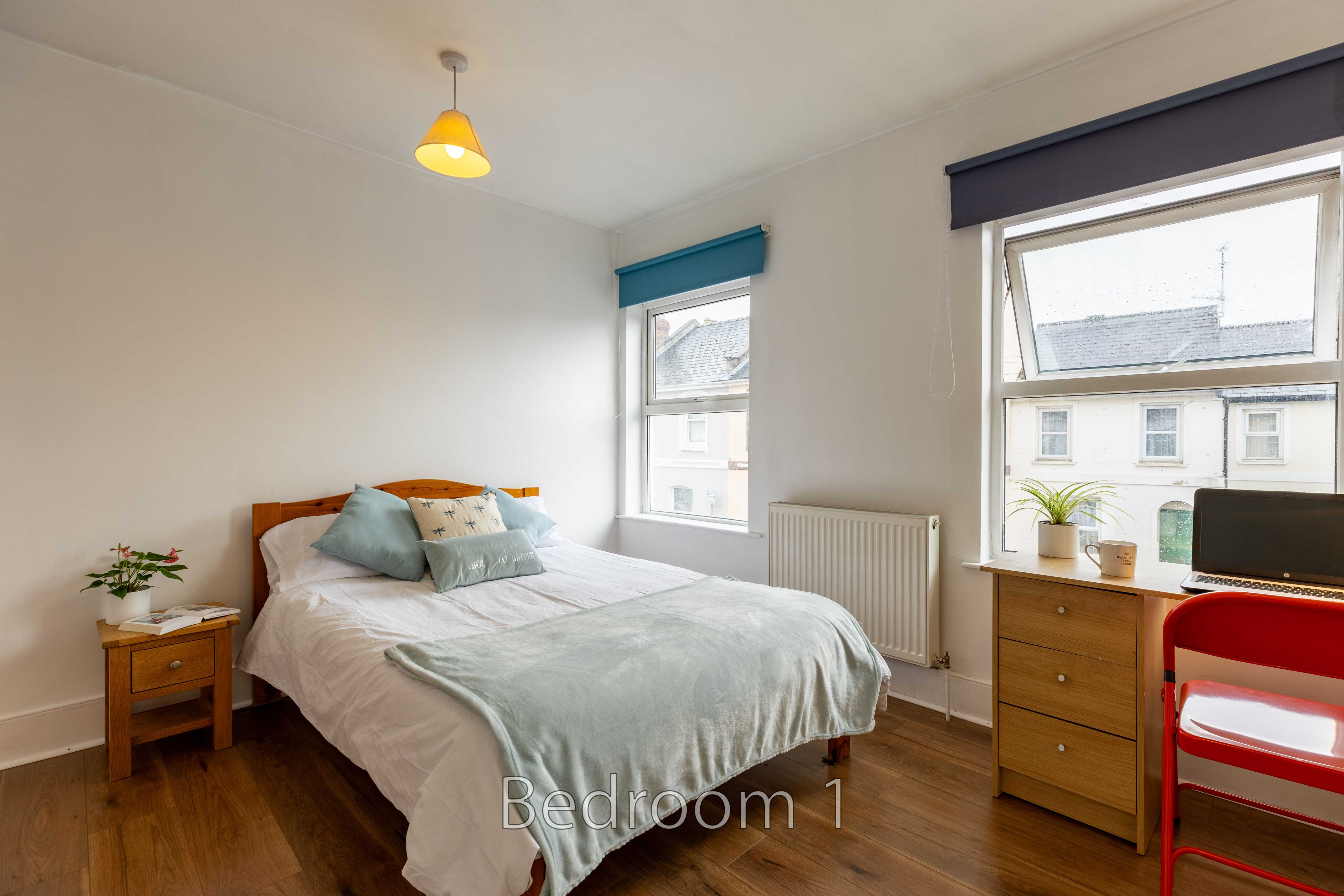 A bedroom with a single bed, bedside table, chest of drawers, desk, and two windows.