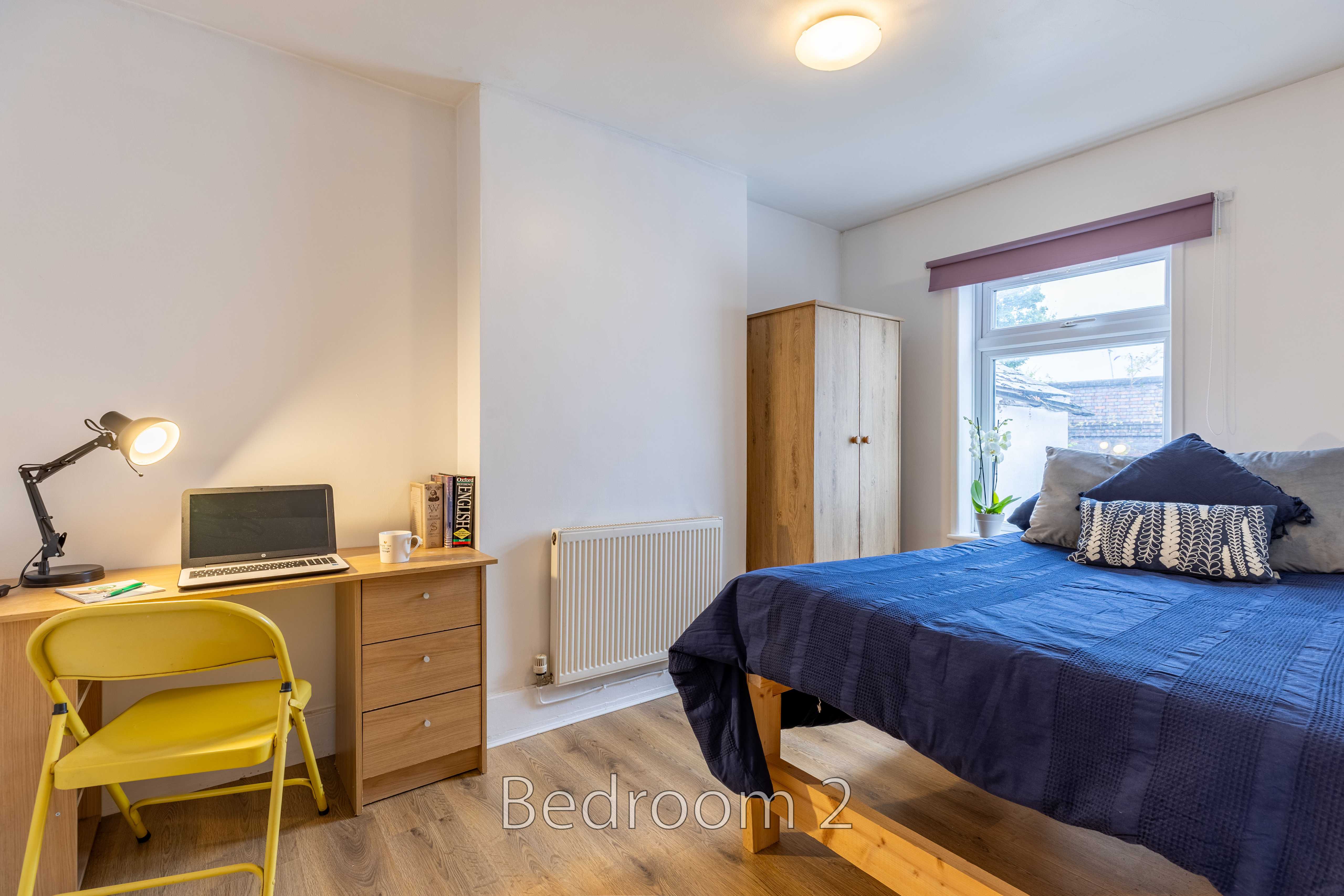 A furnished bedroom with a double bed, desk, chair, wardrobe, radiator and window.