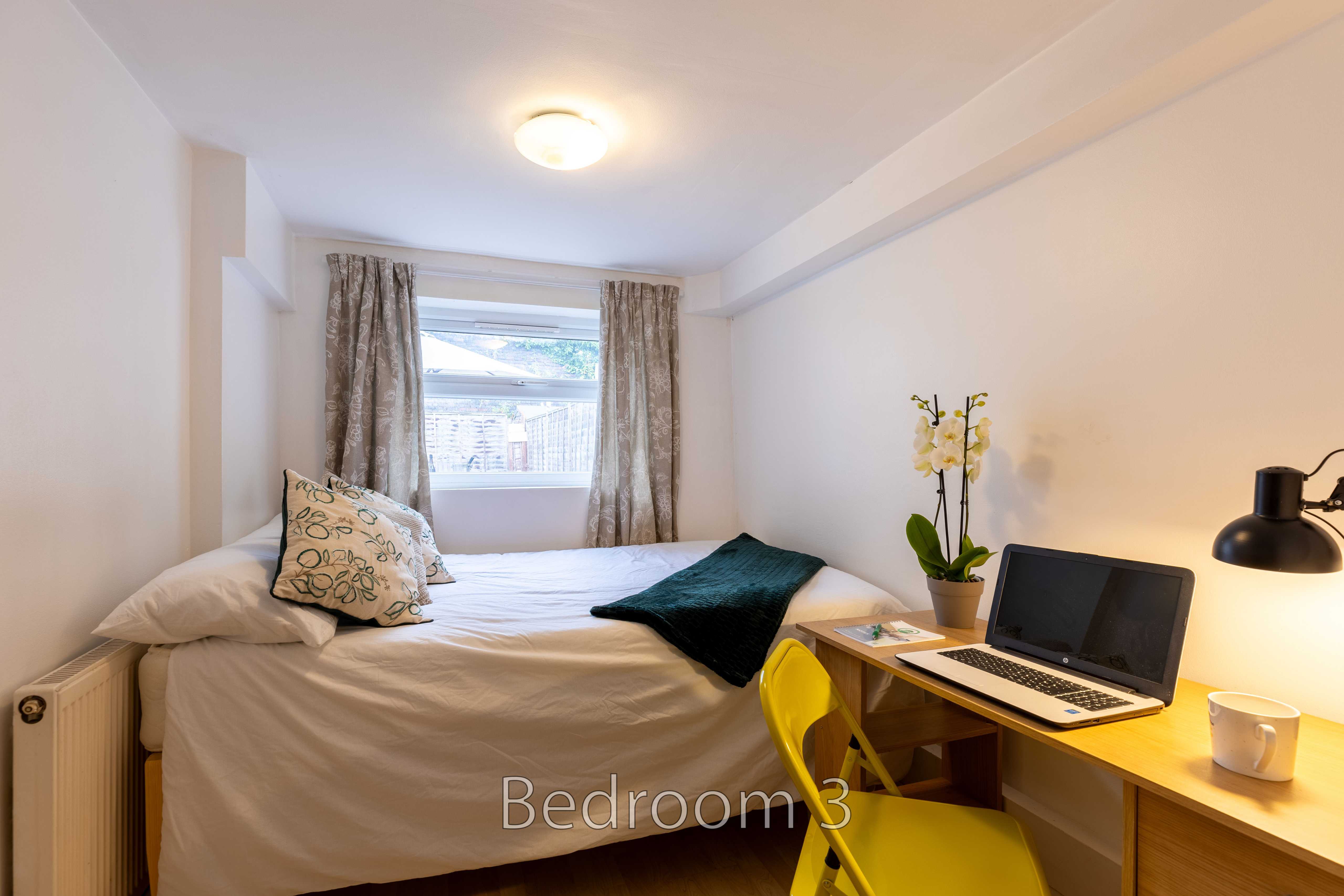 A furnished bedroom with a bed, desk, laptop, chair, window and curtains.