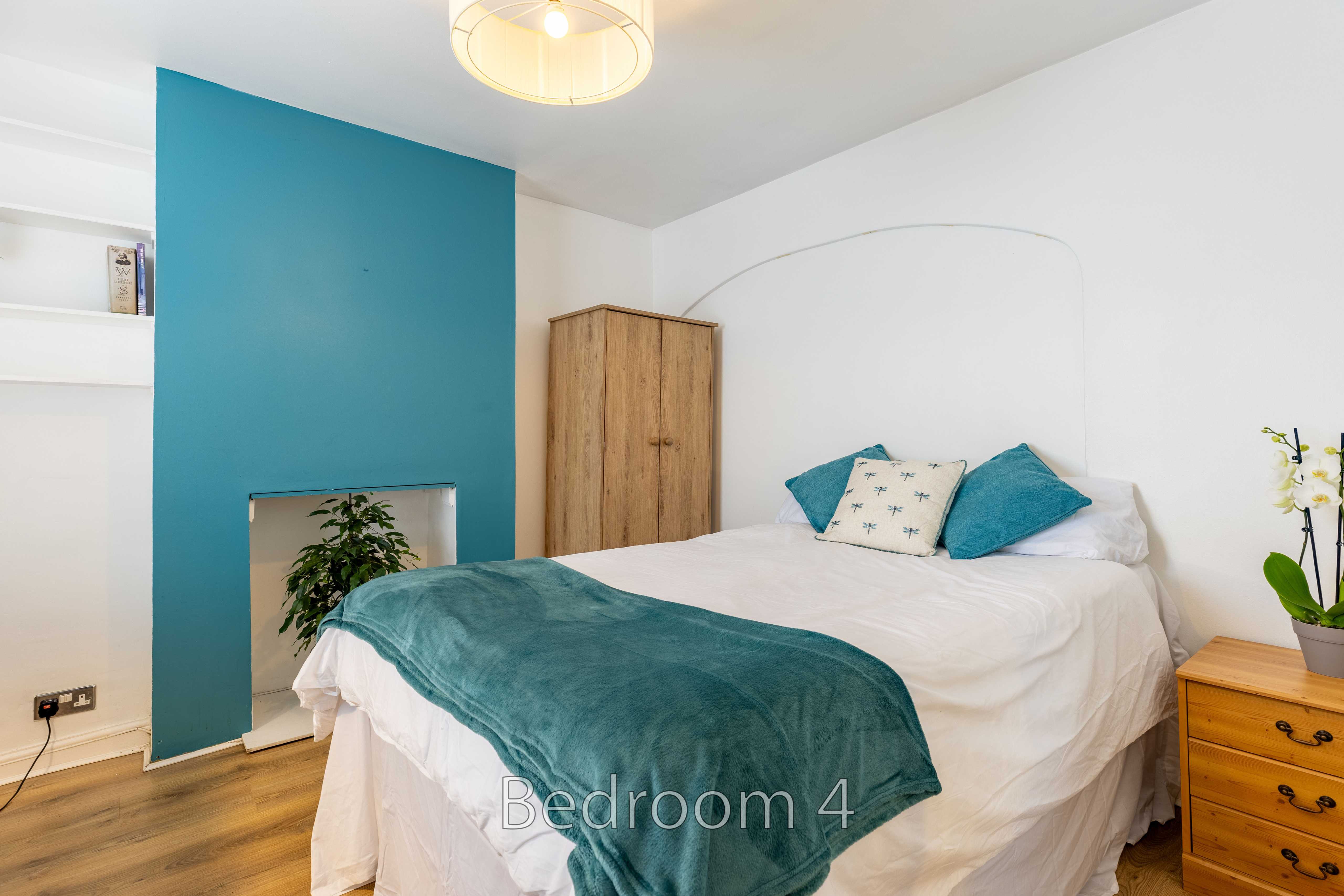 A bedroom with a double bed, teal throw and cushions, wooden wardrobe, chest of drawers, and teal accent wall.