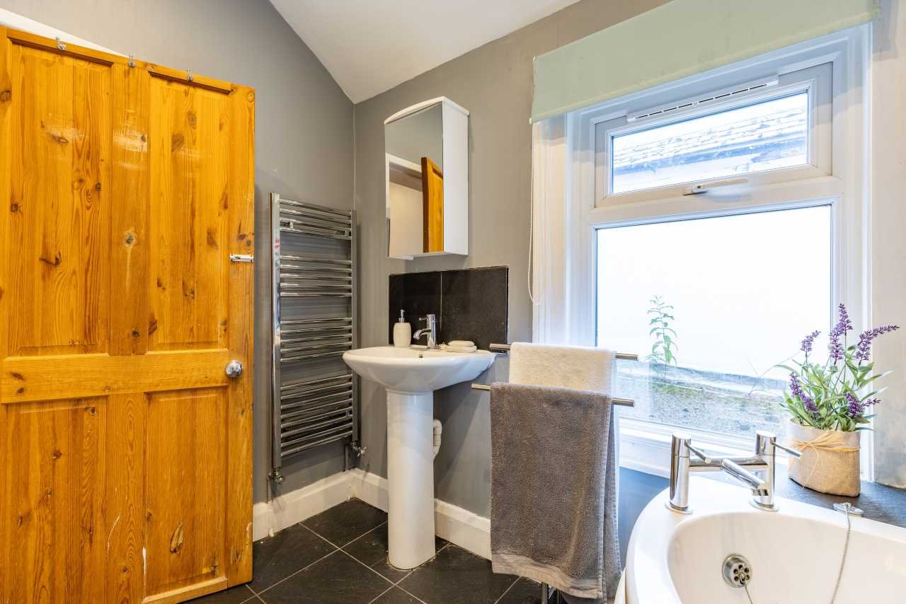 A small bathroom with a wooden door, pedestal sink, towel radiator, bathtub, and window.