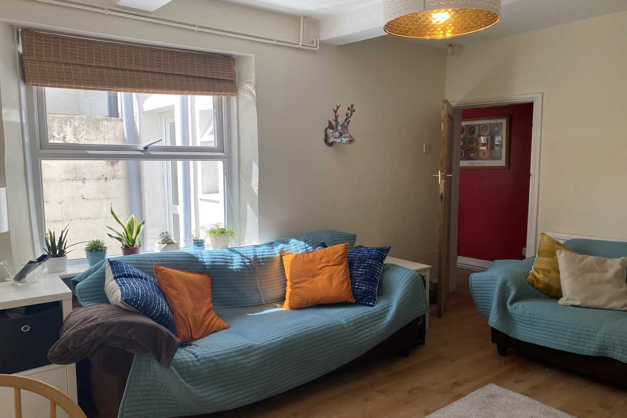 A furnished living room with two blue sofas, colorful cushions, a window, plants, and a wooden floor.
