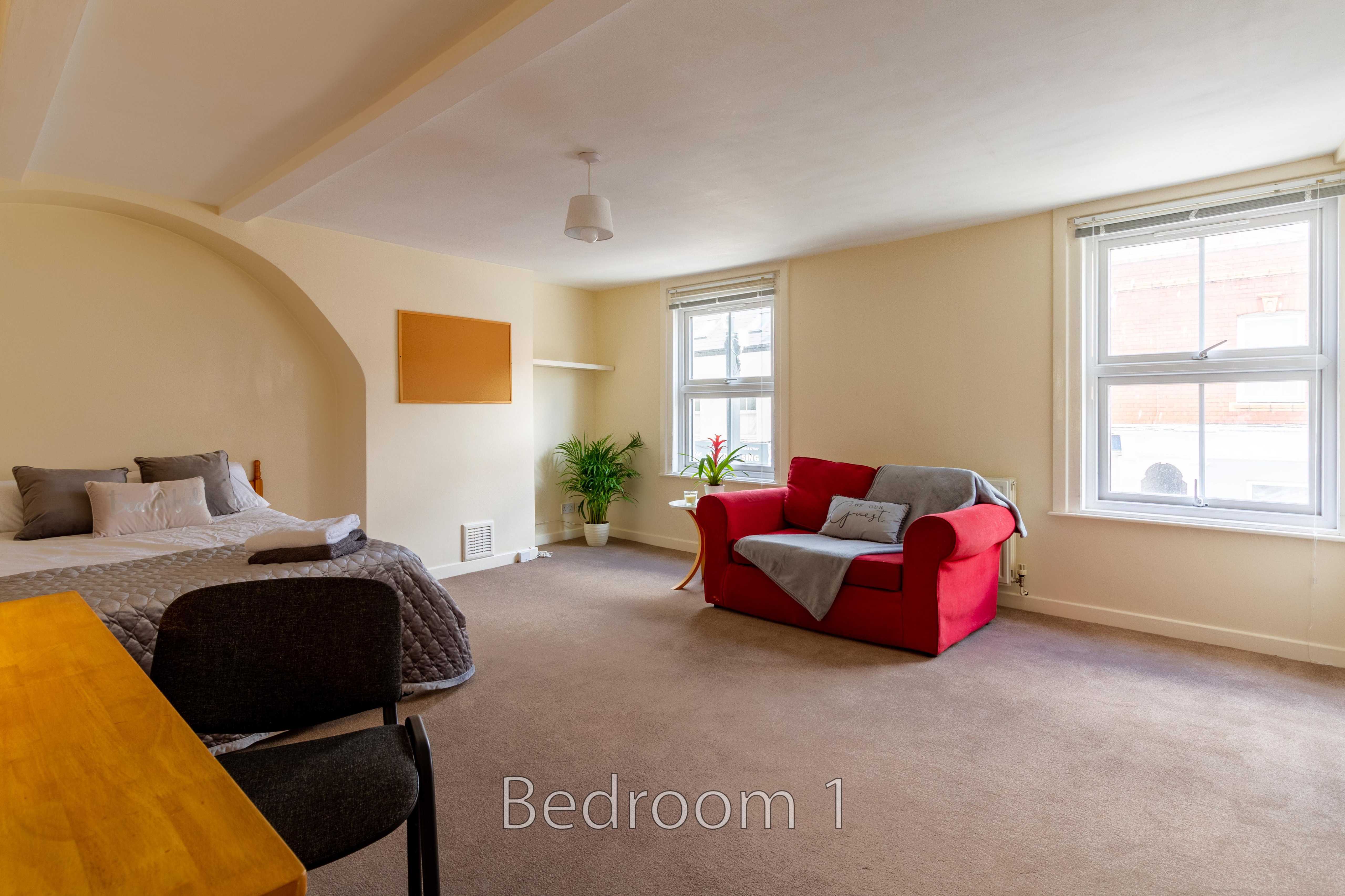 A furnished bedroom with a bed, red armchair, desk, and two windows.