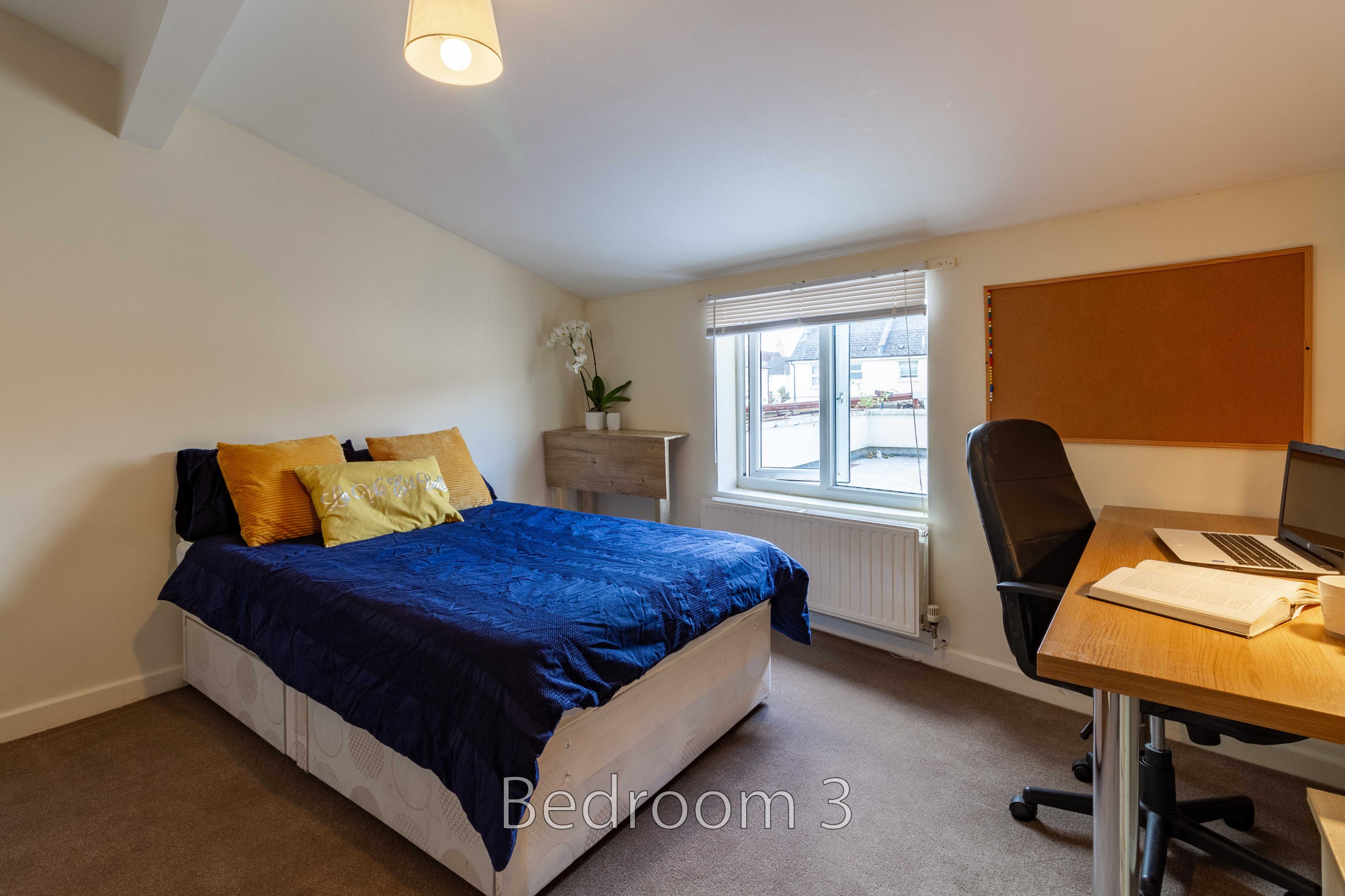 A furnished bedroom with a double bed, desk and chair, window, radiator and chest of drawers.