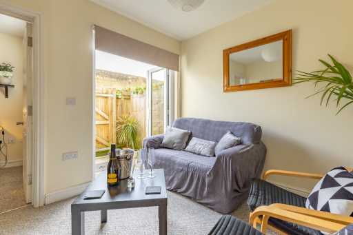 A furnished sitting room with a sofa, coffee table, mirror, armchair, and an open door to a small garden.